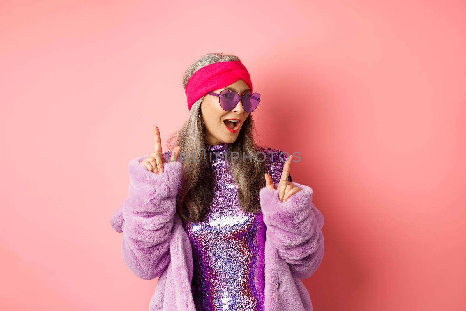Hipster granny in sunglasses and glittering dress posing for photo with peace, victory signs, standing sassy against pink background.
