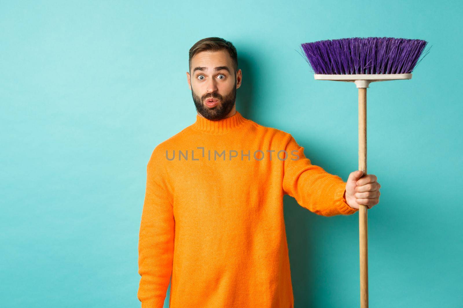 Shocked man receiving broom to do house chores, looking confused, standing over light blue background.