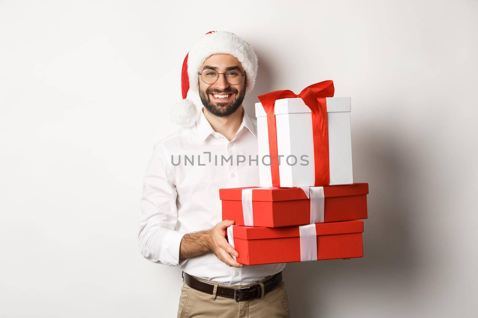 Merry christmas, holidays concept. Happy young man smiling, holding gifts in boxes and wearing santa hat, white background by Benzoix