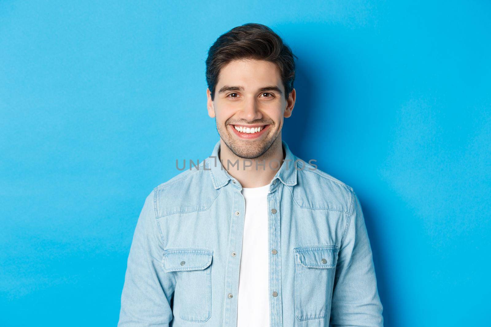 Close-up of young successful man smiling at camera, standing in casual outfit against blue background.