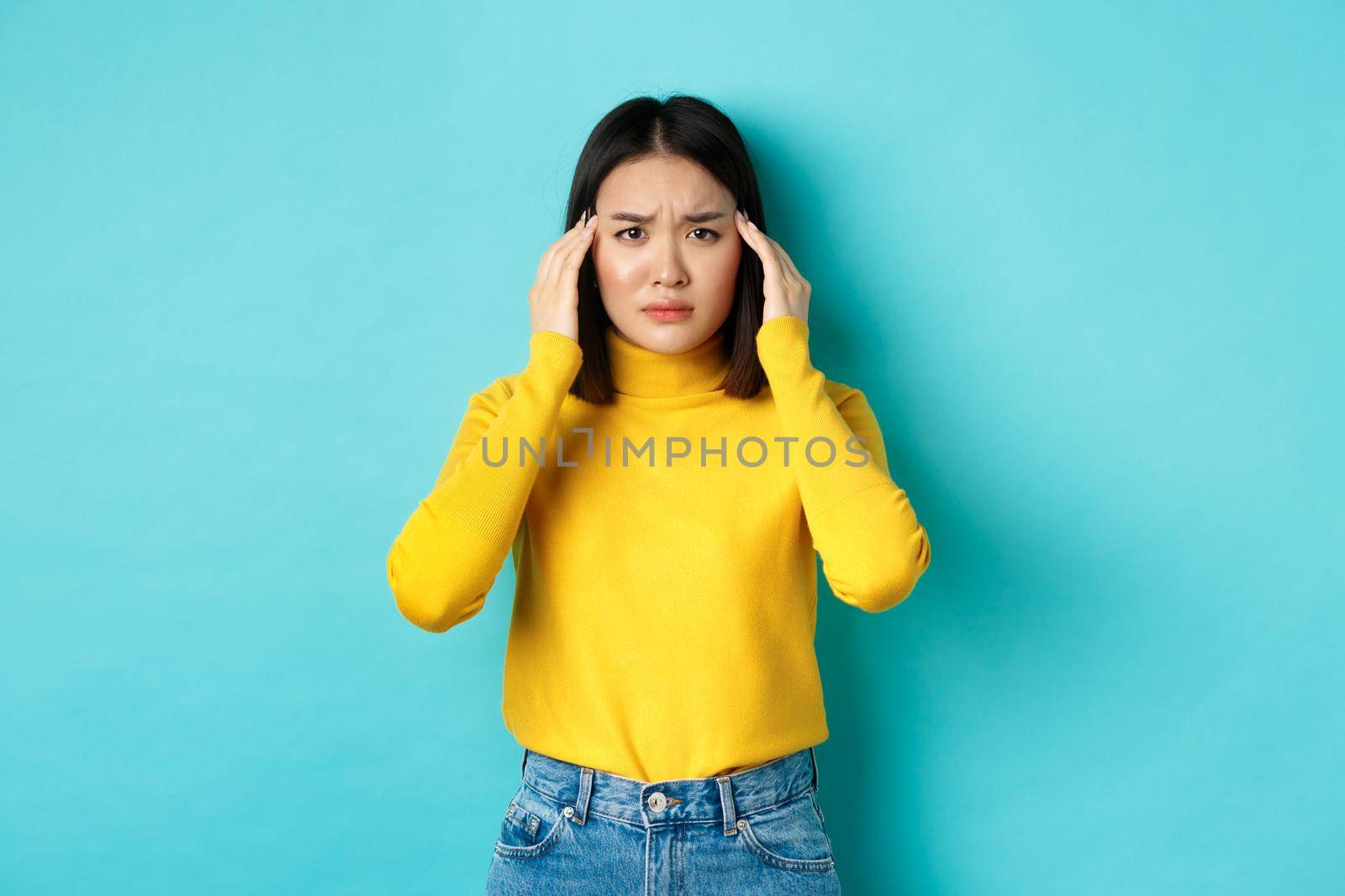 Image of distressed asian woman touching head and frowning, feeling headache, standing troubled against blue background.