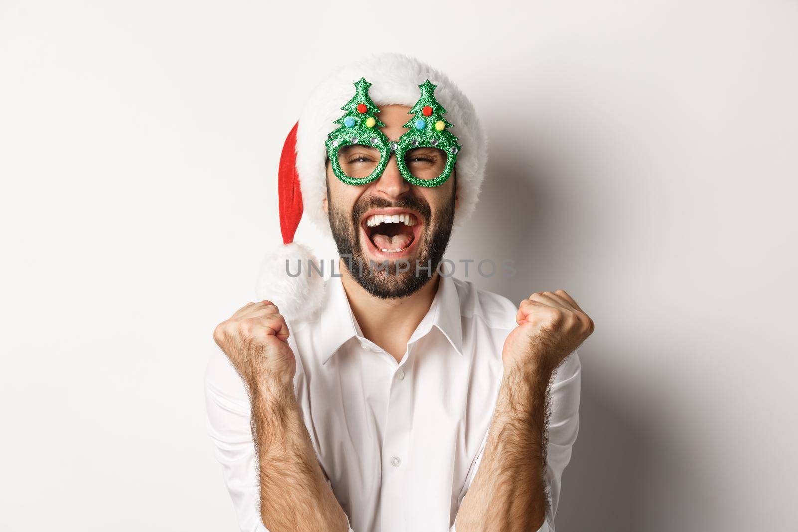 Close-up of man celebrating christmas or new year, wearing xmas party glasses and santa hat, rejoicing and shouting of joy, standing over white background by Benzoix