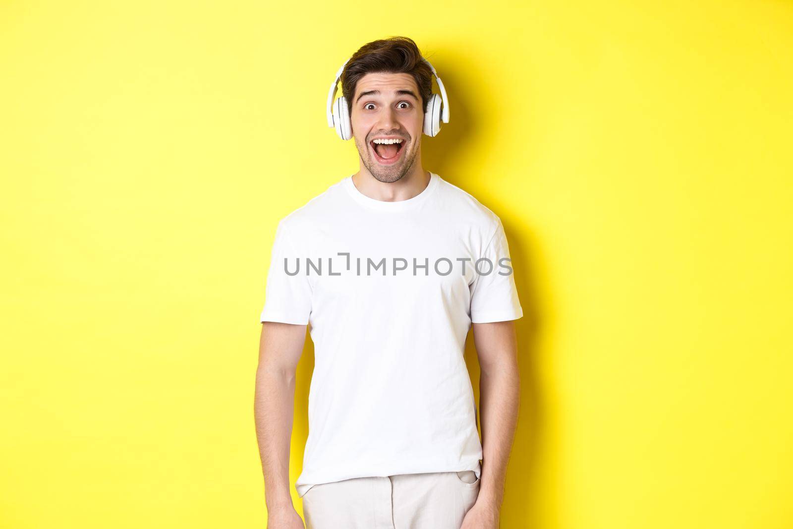 Man in headphones looking surprised, standing against yellow background in white outfit.