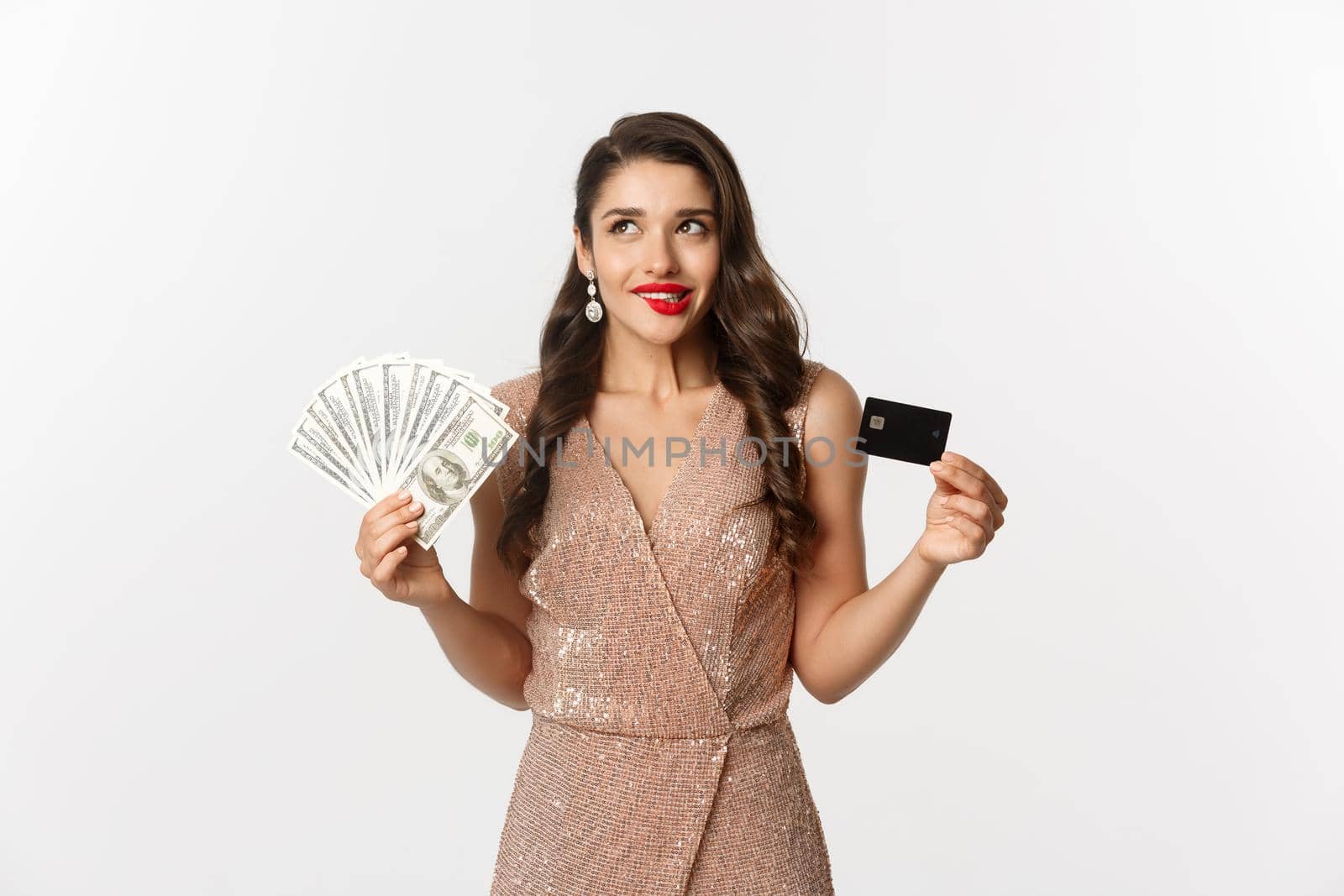Shopping concept. Young woman deciding what to buy, looking thoughtful at upper left corner and showing credit card with money, standing in party dress by Benzoix