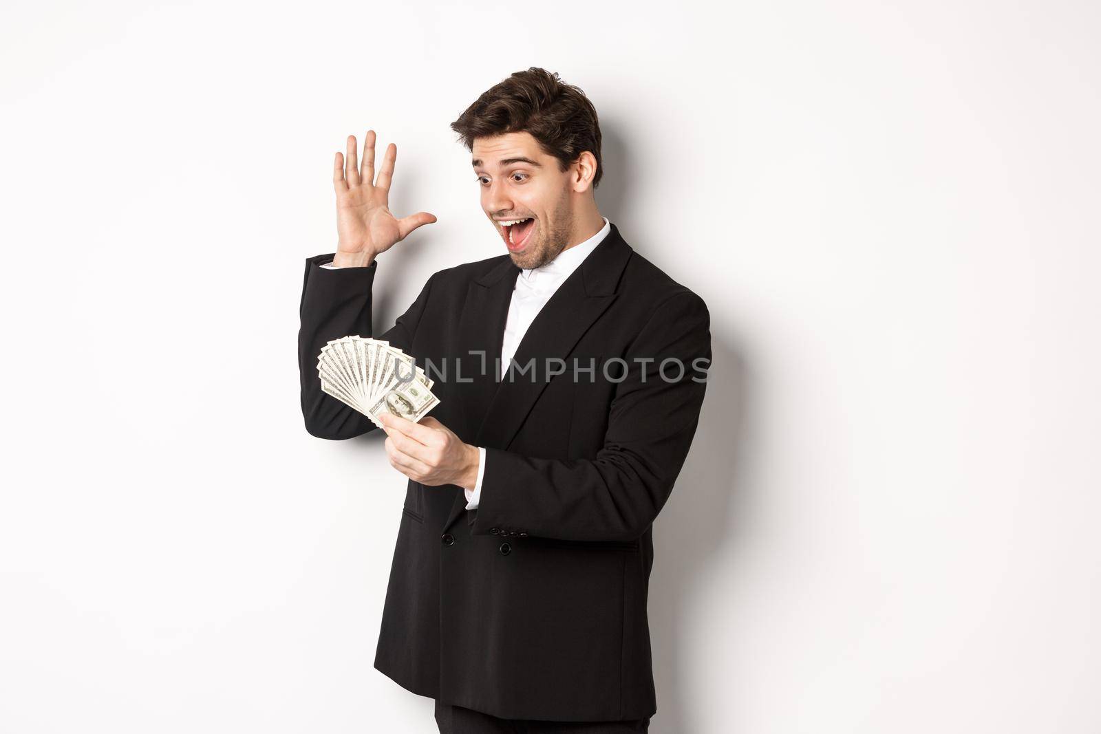 Image of successful businessman in black suit, looking at money and triumphing, earn cash, standing over white background.