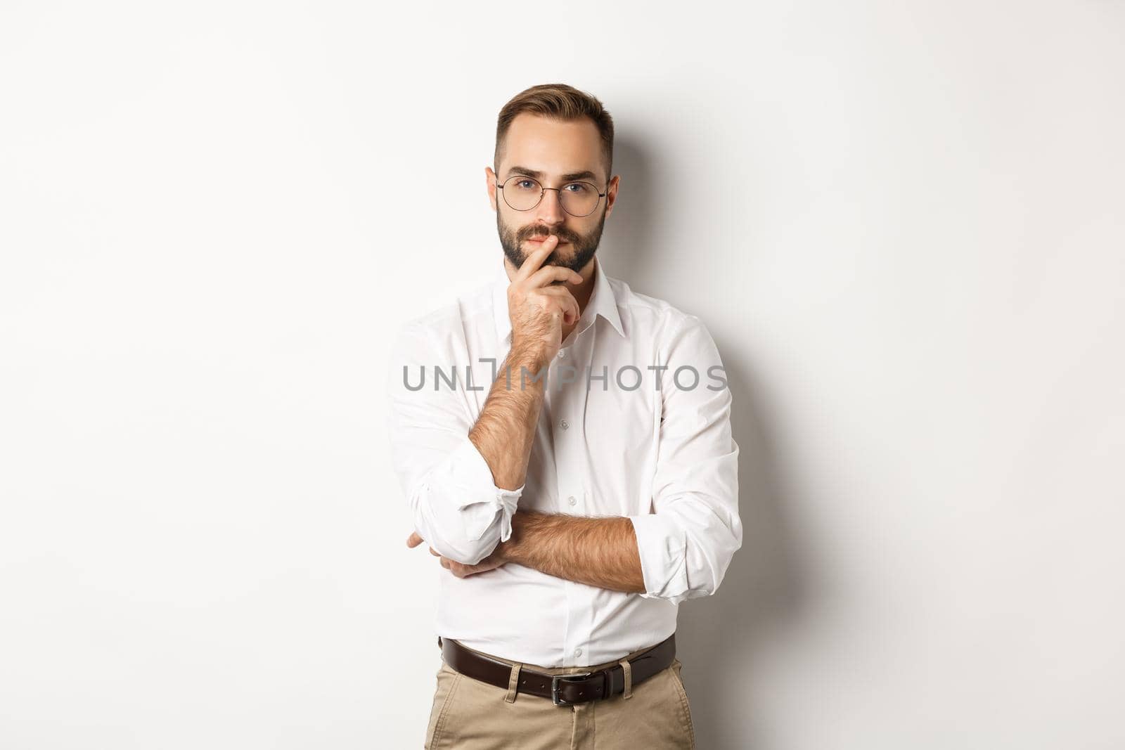 Thoughtful handsome businessman looking at camera, making choice or thinking, standing in glasses and white collar shirt against studio background by Benzoix