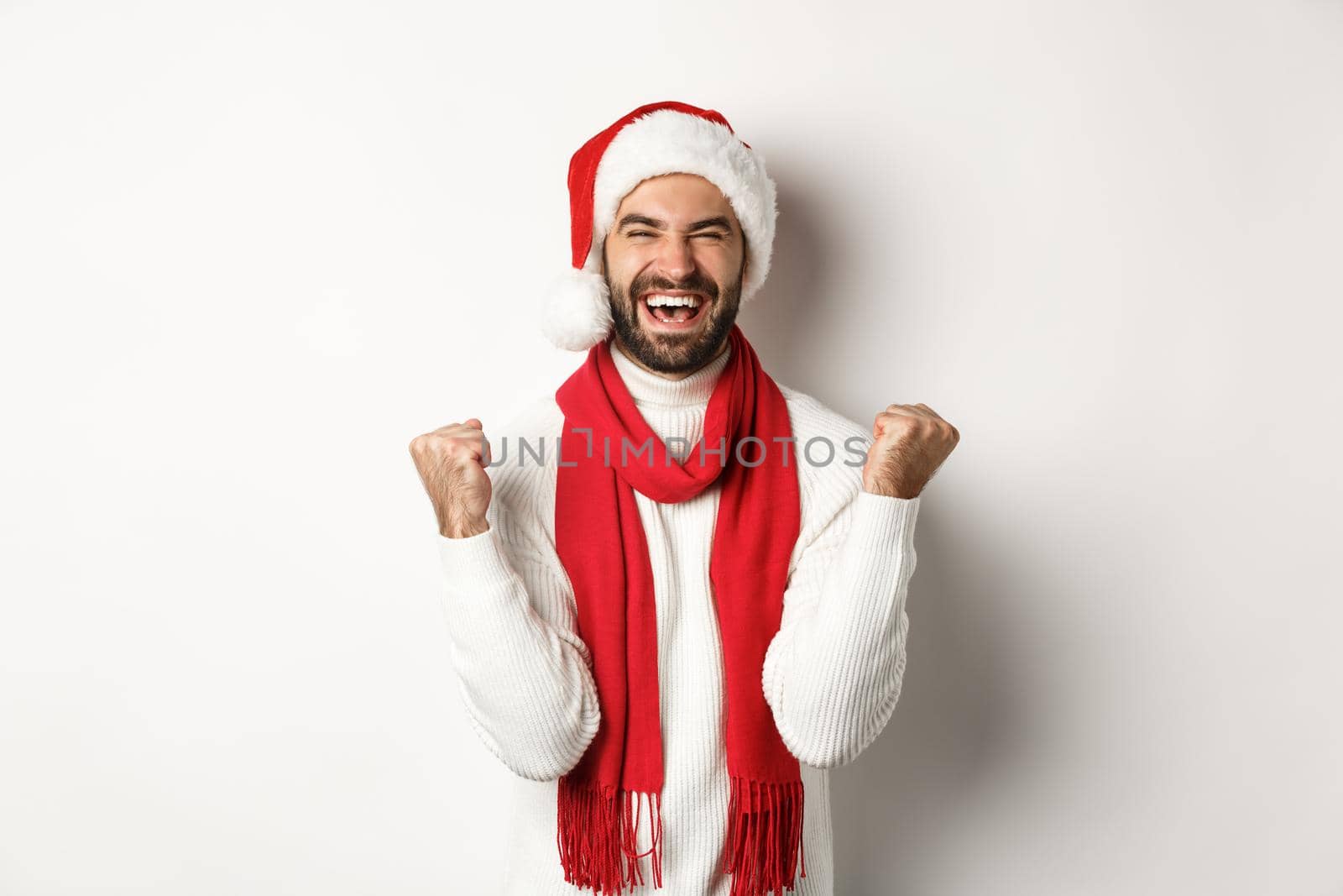 Christmas holidays. Man winner celebrating and triumphing, raising hands up in rejoice, achieve goal, wearing Santa hat and red scarf, white background.