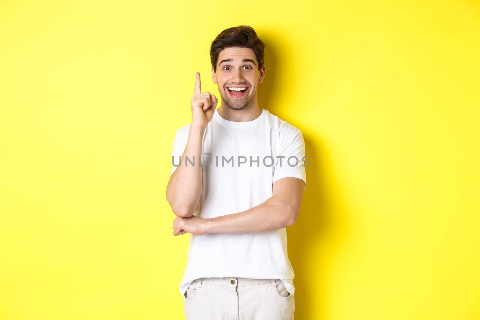Image of attractive guy having an idea, raising finger up and suggesting plan, smiling excited, standing over yellow background.