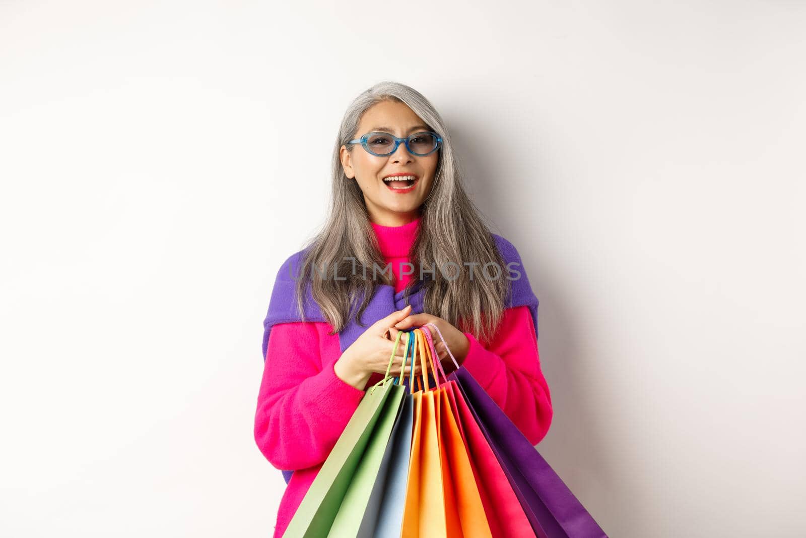 Stylish senior asian woman in sunglasses going shopping on holiday sale, holding paper bags and smiling, standing over white background by Benzoix