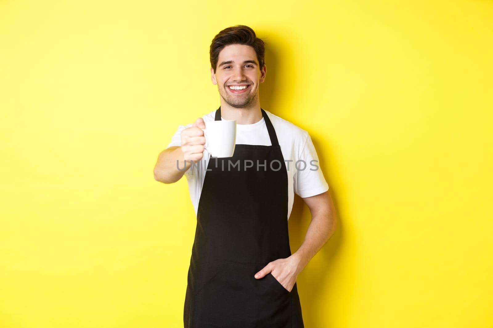 Handsome barista in black apron giving you cup of coffee and smiling, standing over yellow background.