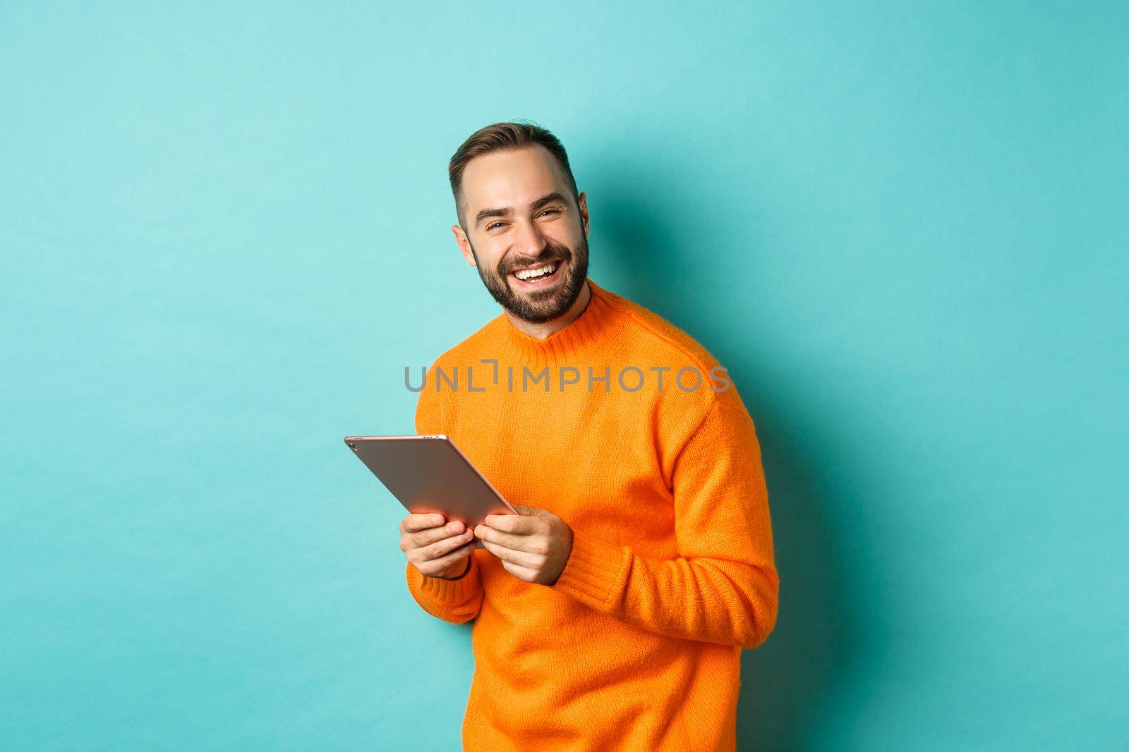 Handsome bearded man using digital tablet, laughing at camera, standing happy against light blue background.