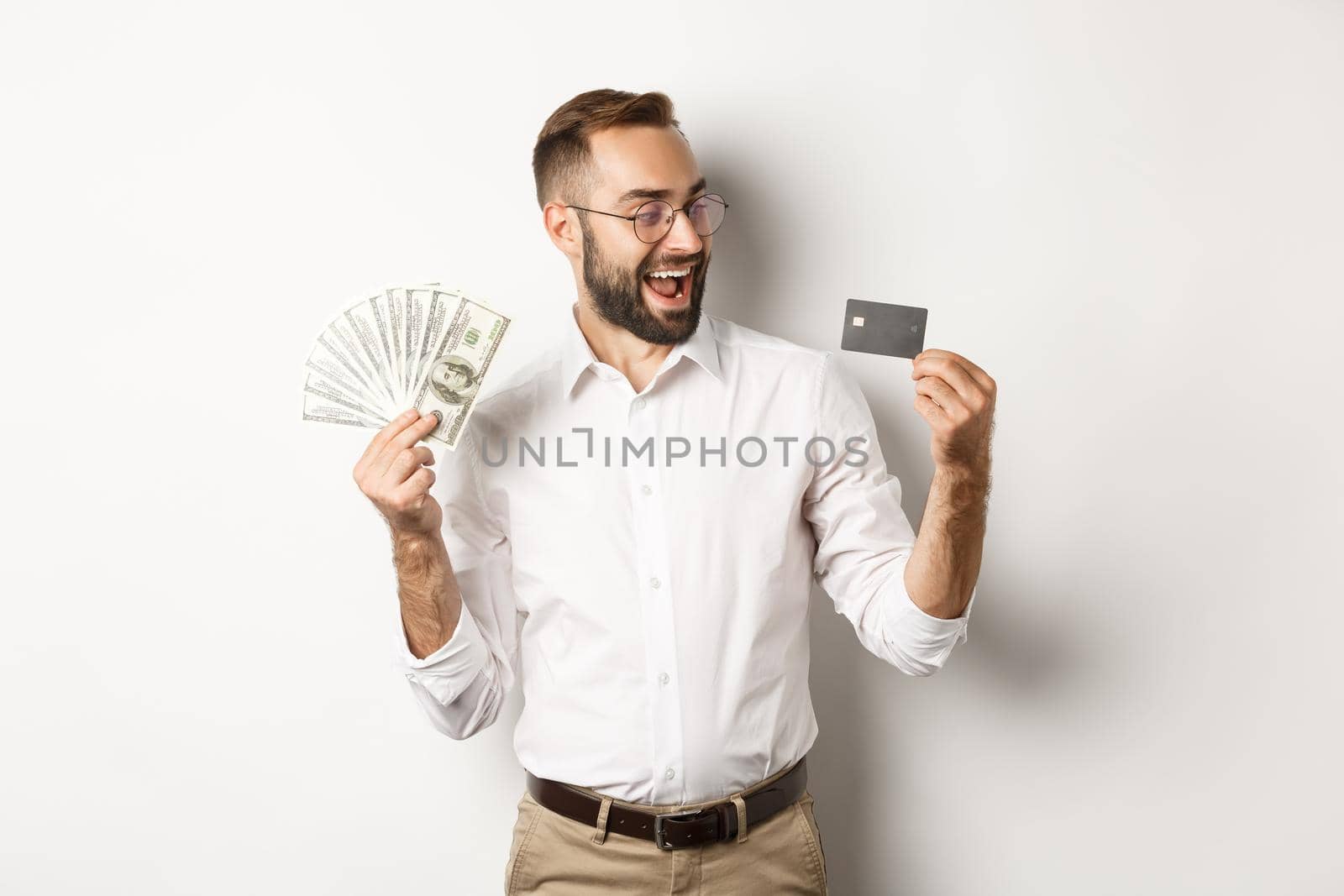 Excited businessman holding money and looking at credit card, standing over white background.