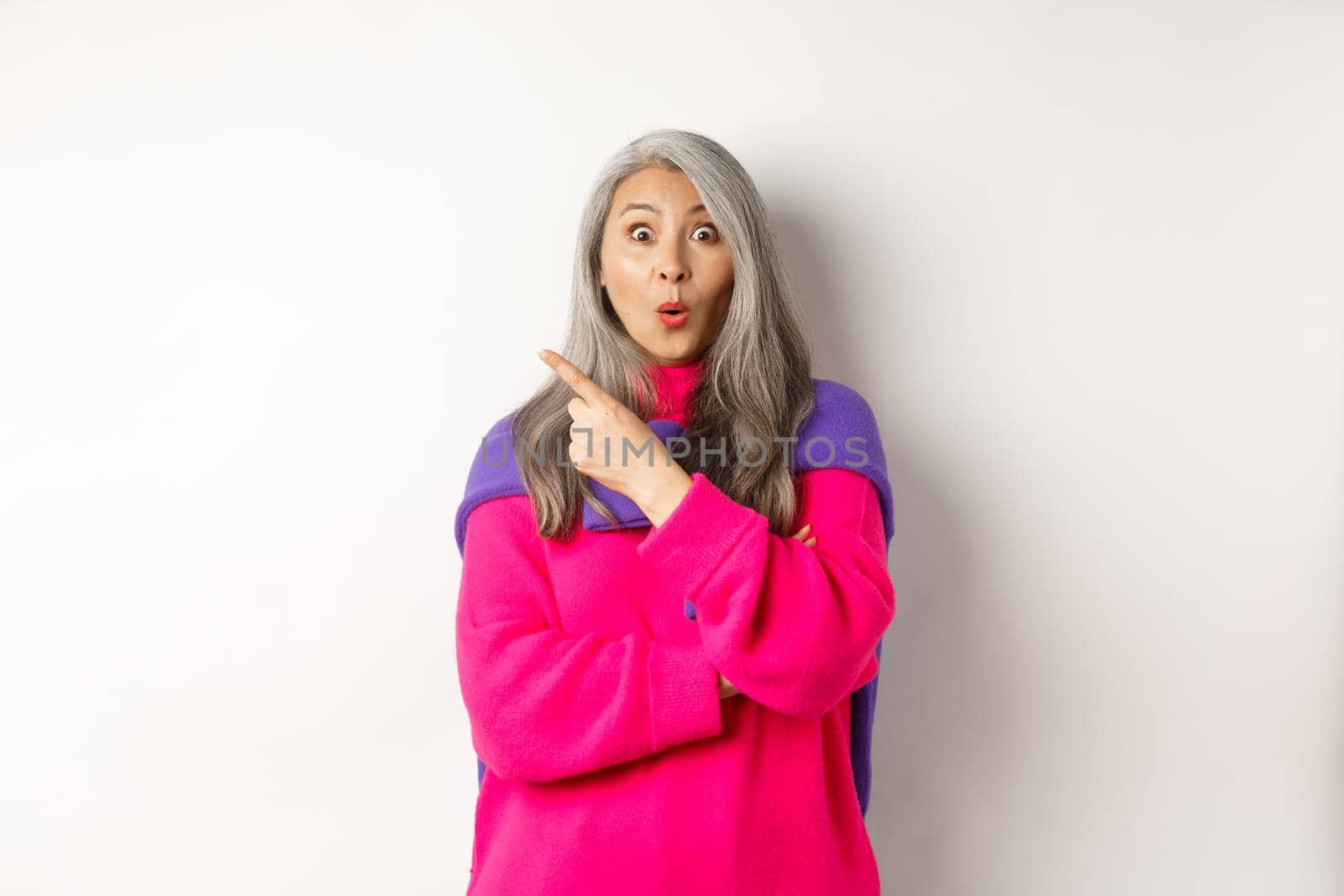 Portrait of impressed asian lady checking out promo offer, pointing at upper left corner and staring at camera amazed, standing in trendy pink sweater over white background.