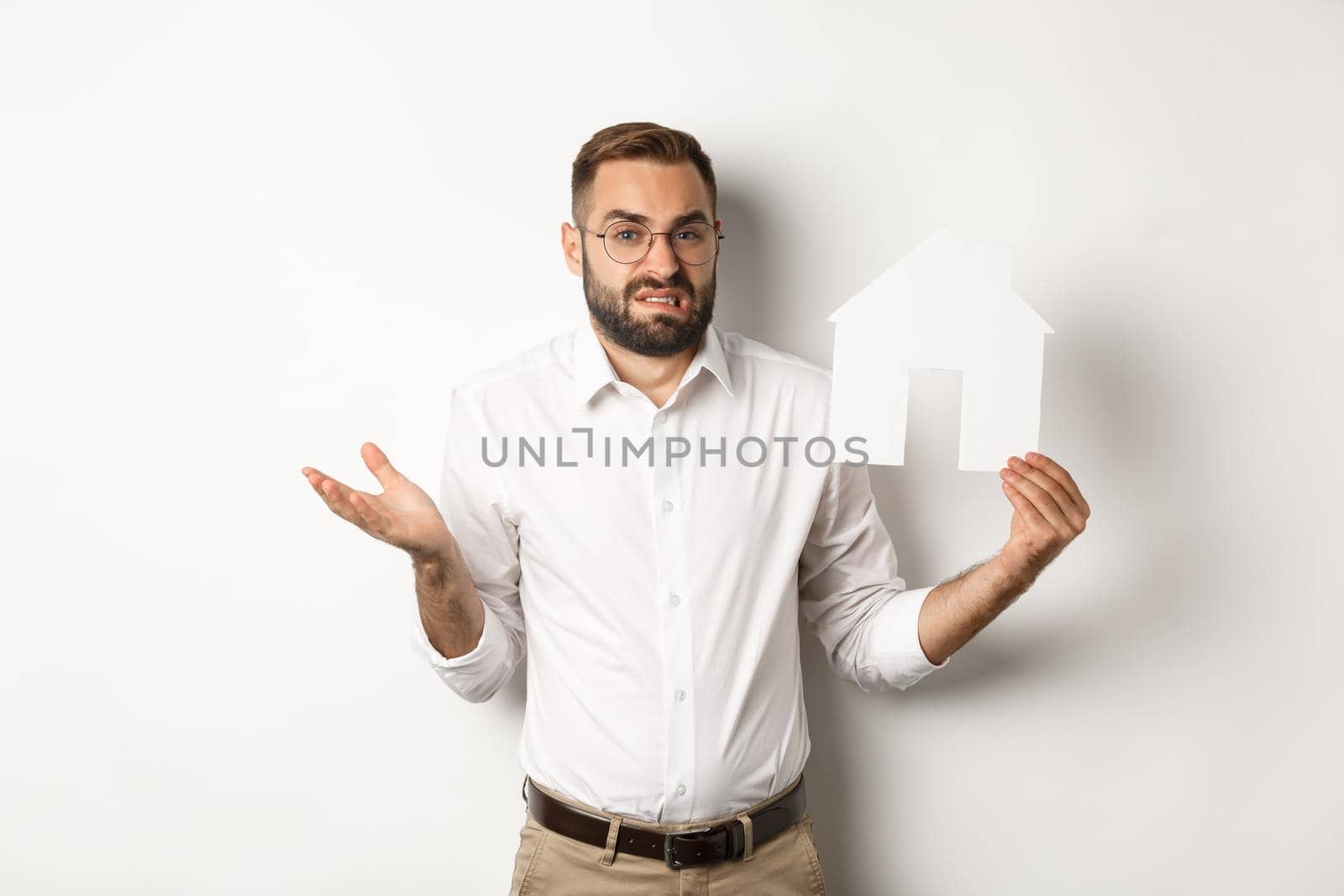 Real estate. Confused man shrugging, showing house paper model and looking indecisive, standing over white background by Benzoix