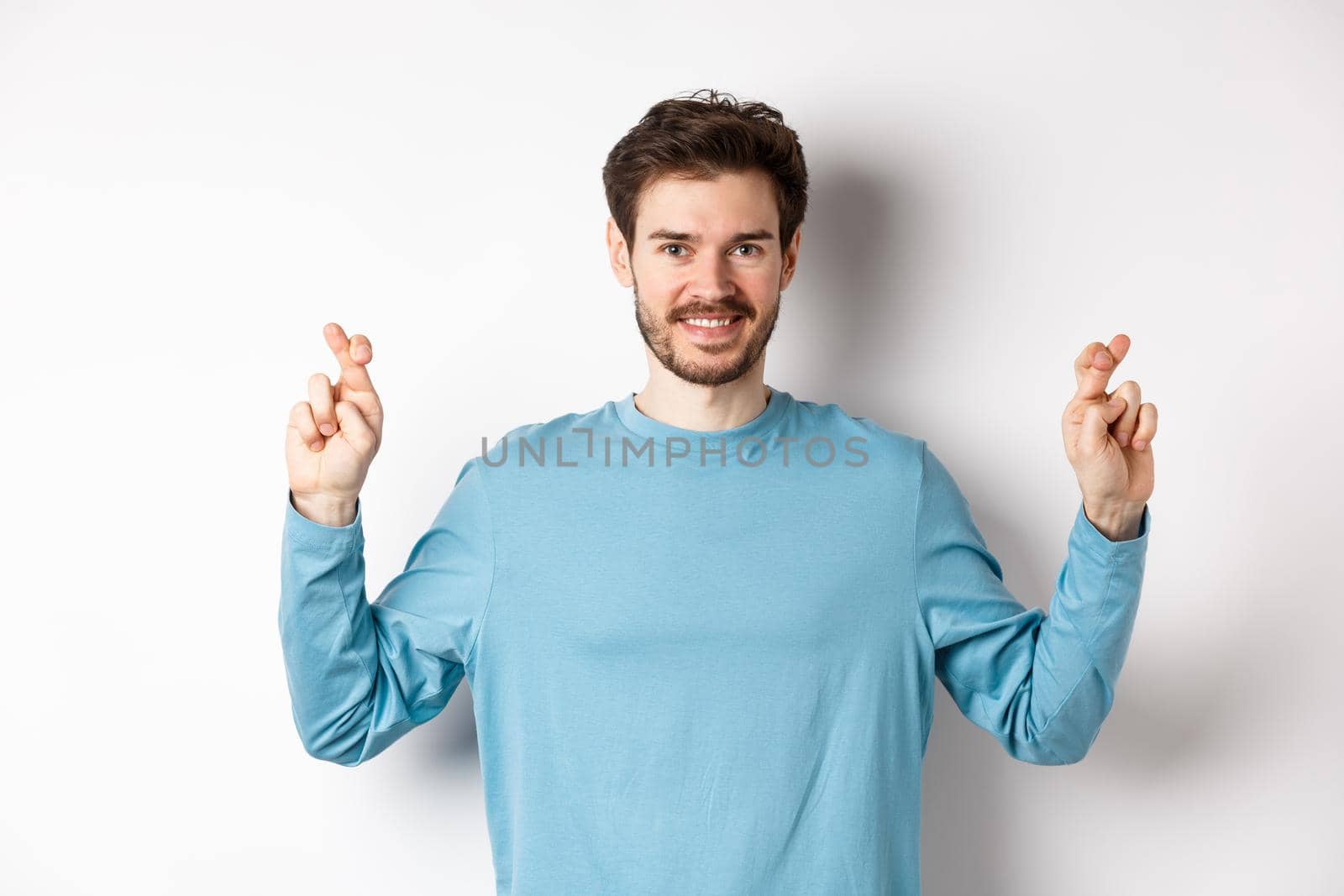 Optimistic smiling man cross fingers for good luck, making wish and waiting for results, standing over white background.