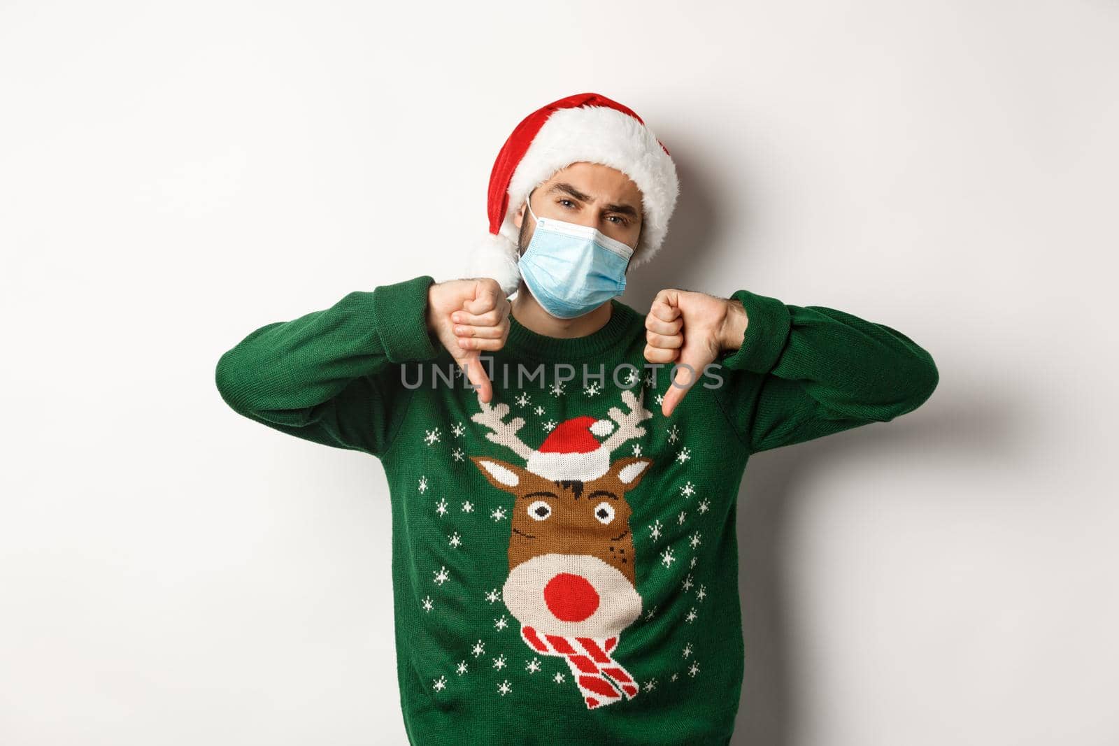 Concept of covid-19 and Christmas holidays. Displeased guy in face mask and santa hat showing thumbs down, standing over white background by Benzoix