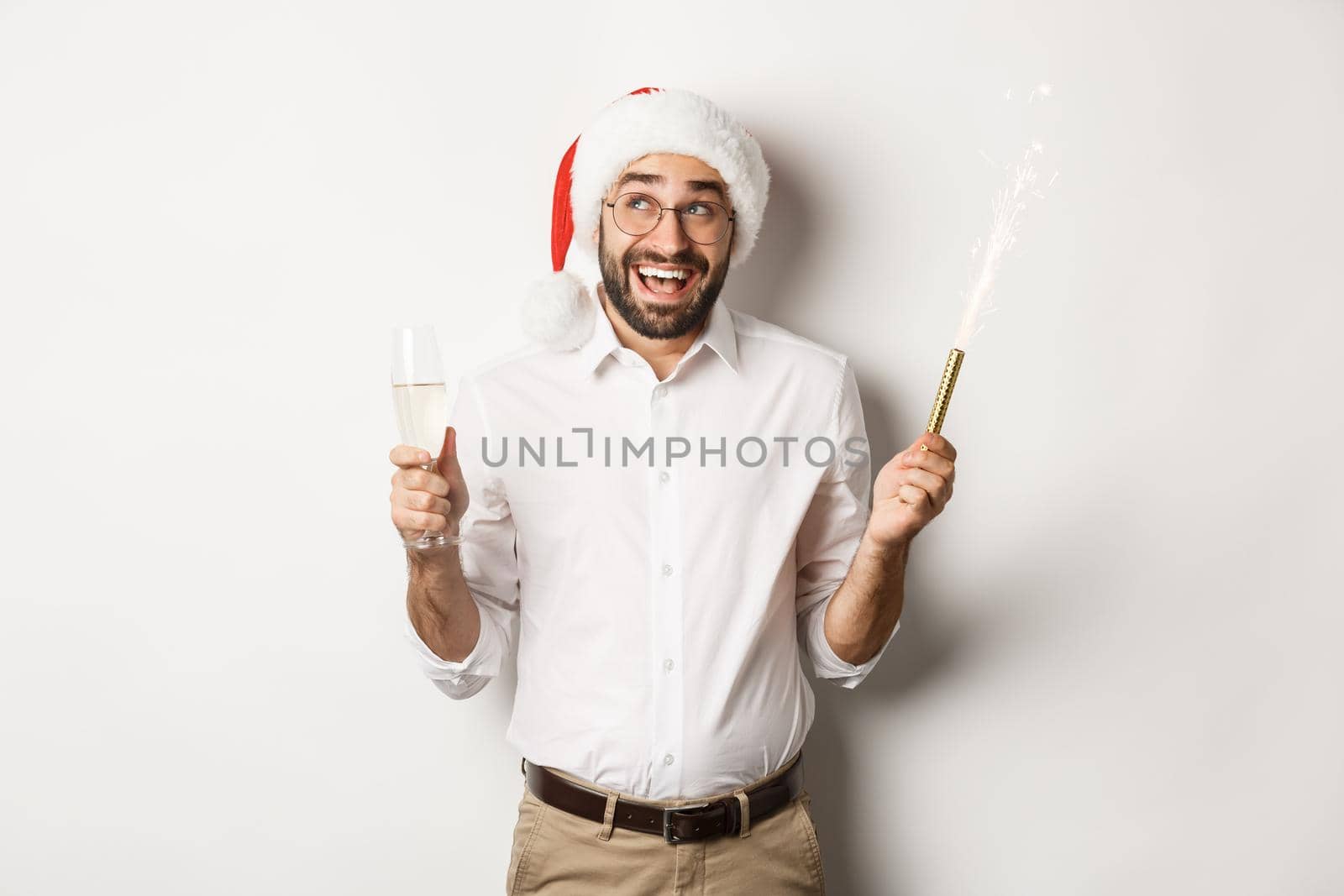 Winter holidays and celebration. Happy businessman enjoying New Year party, wearing Santa hat and drinking champagne, smiling amused, white background.