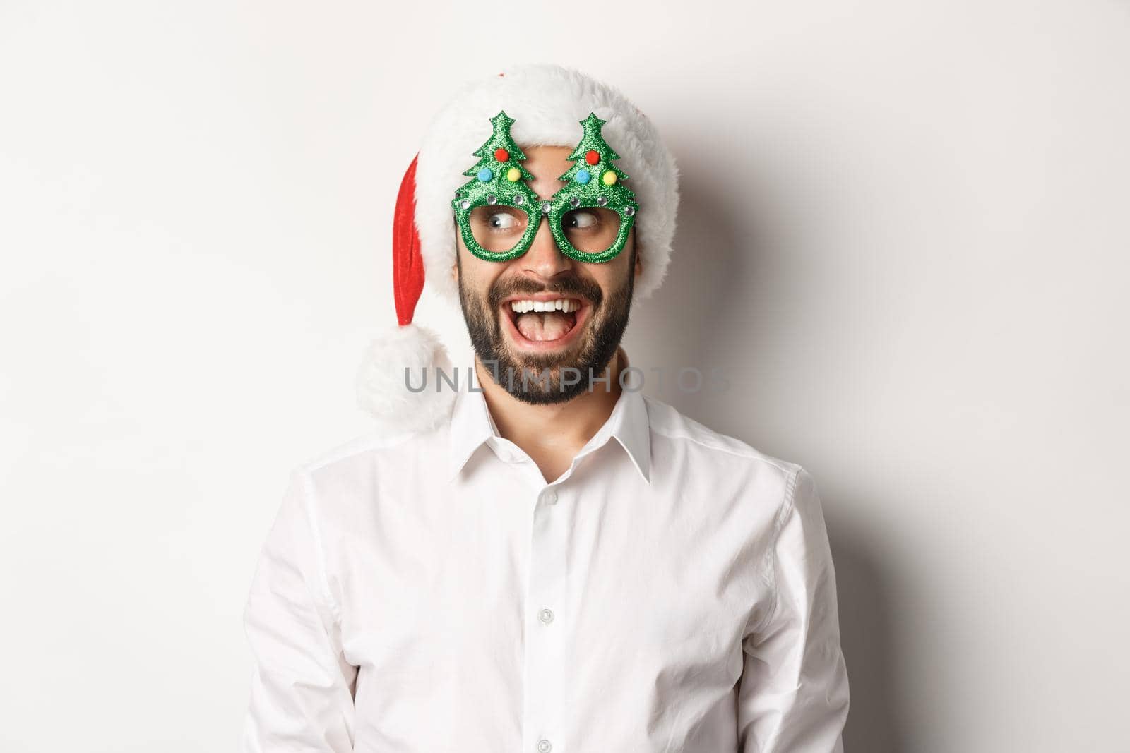 Close-up of funny man looking left with surprised face, wearing christmas party glasses and santa hat, celebrating New Year, white background.