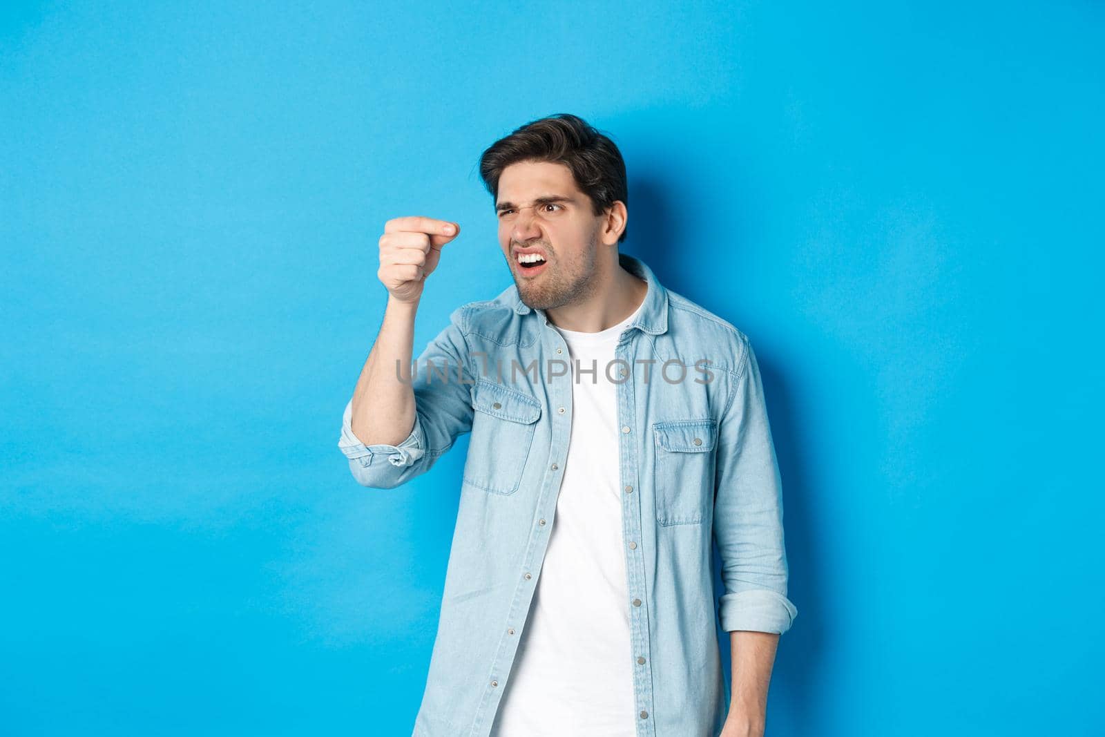 Confused guy holding something tiny in fingers and looking at it puzzled, standing over blue background.
