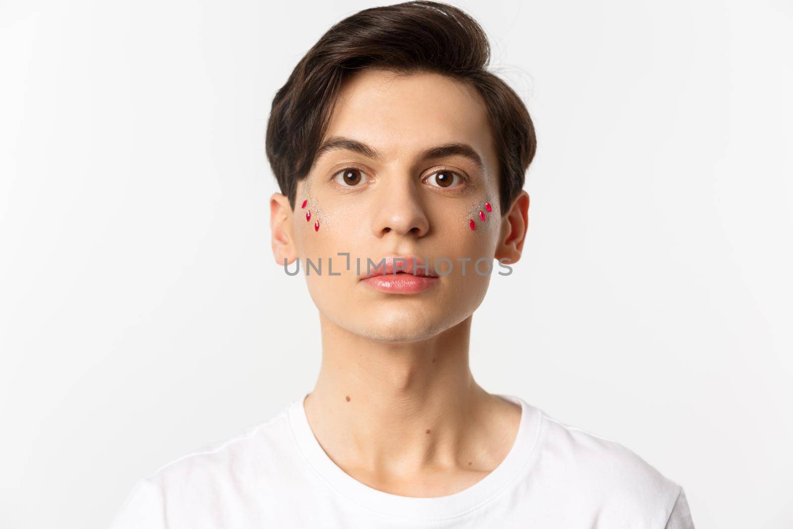 People, lgbtq and beauty concept. Close-up of beautiful androgynous man with glitter on face, looking at camera, standing over white background.