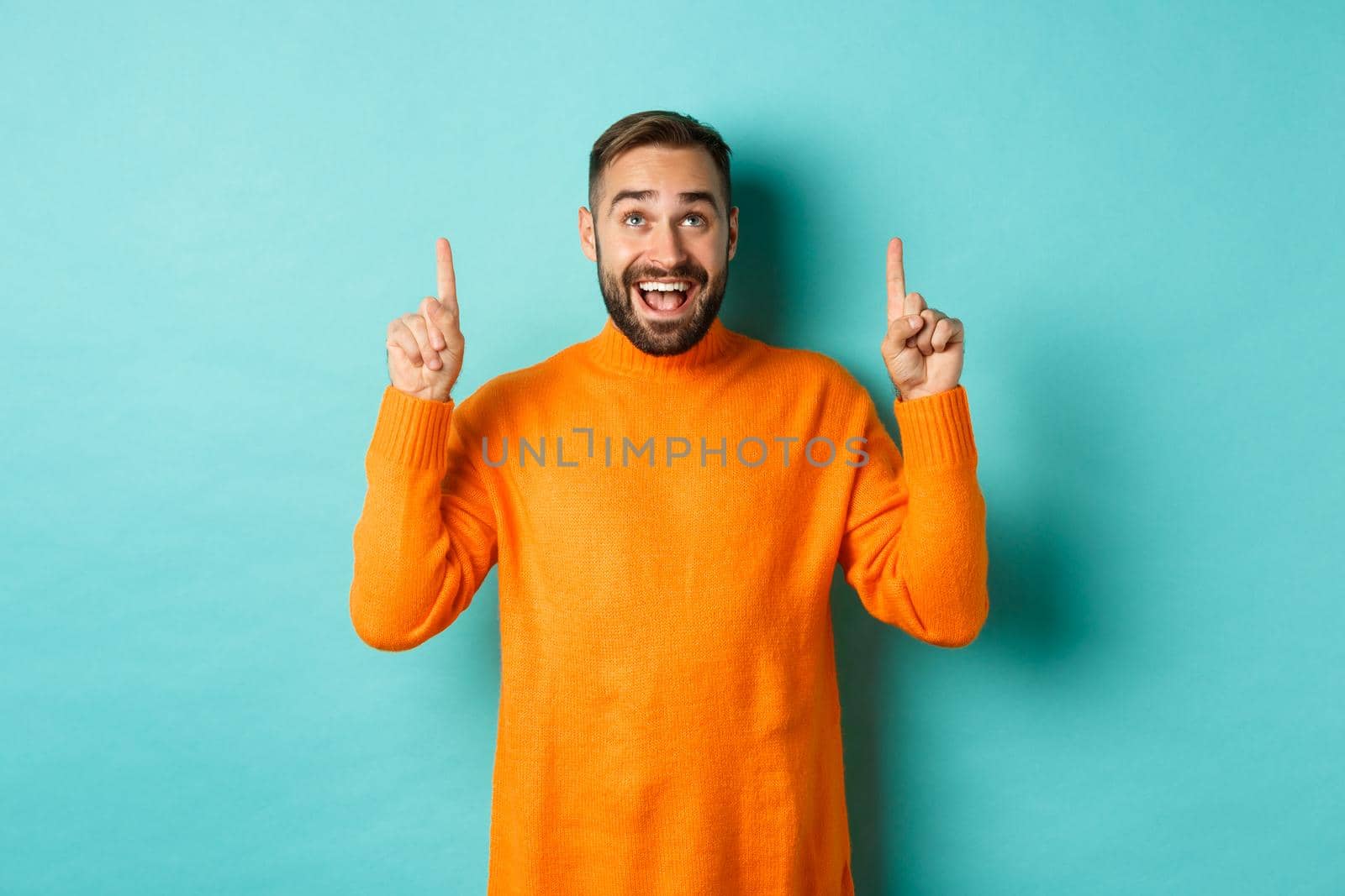 Happy caucasian man showing winter holidays promo offer, pointing fingers up, demonstrate advertisement, standing in sweater against turquoise background by Benzoix
