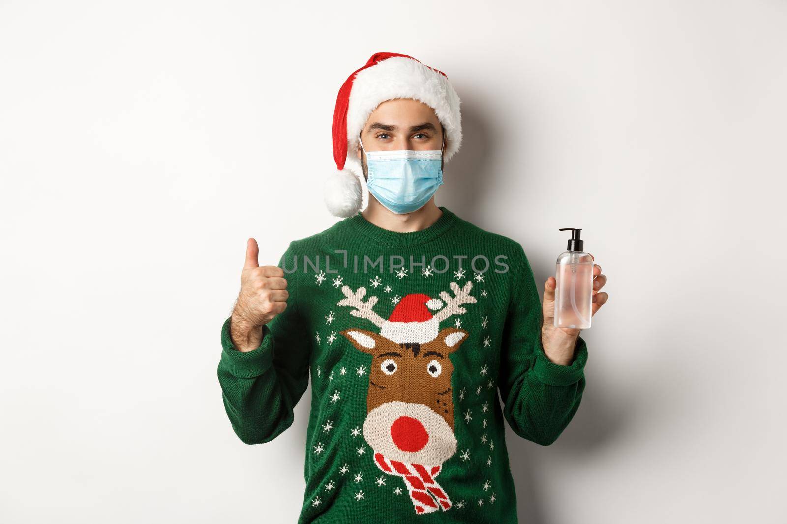 Concept of covid-19 and Christmas holidays. Man recommending hand sanitizer, showing thumb up and antiseptic, wearing medical mask with santa hat, white background by Benzoix