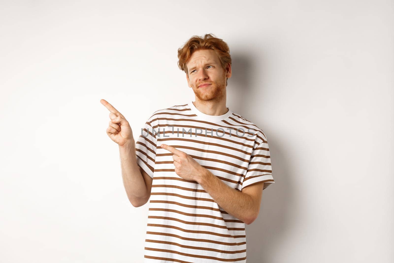 Doubtful young man with bristle and red hair looking, pointing fingers left at something average, staring skeptical at promo, white background by Benzoix