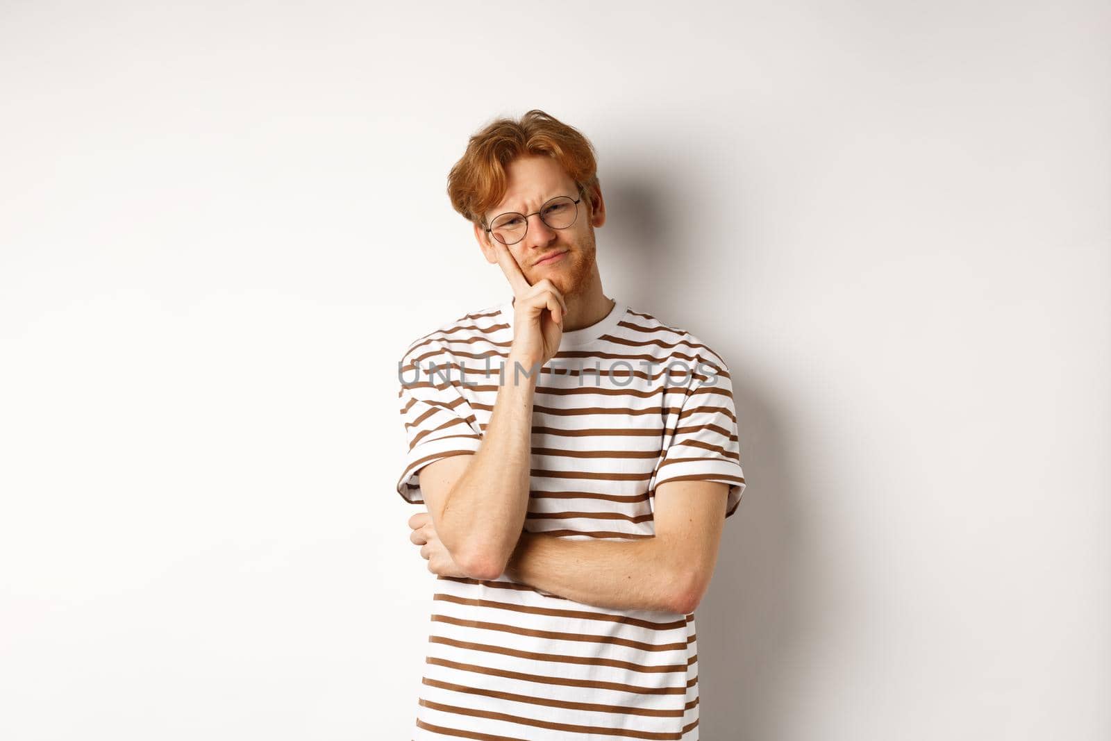 Young man with red messy hair and glasses looking doubtful, thinking and squinting at camera, standing thoughtful over white background by Benzoix