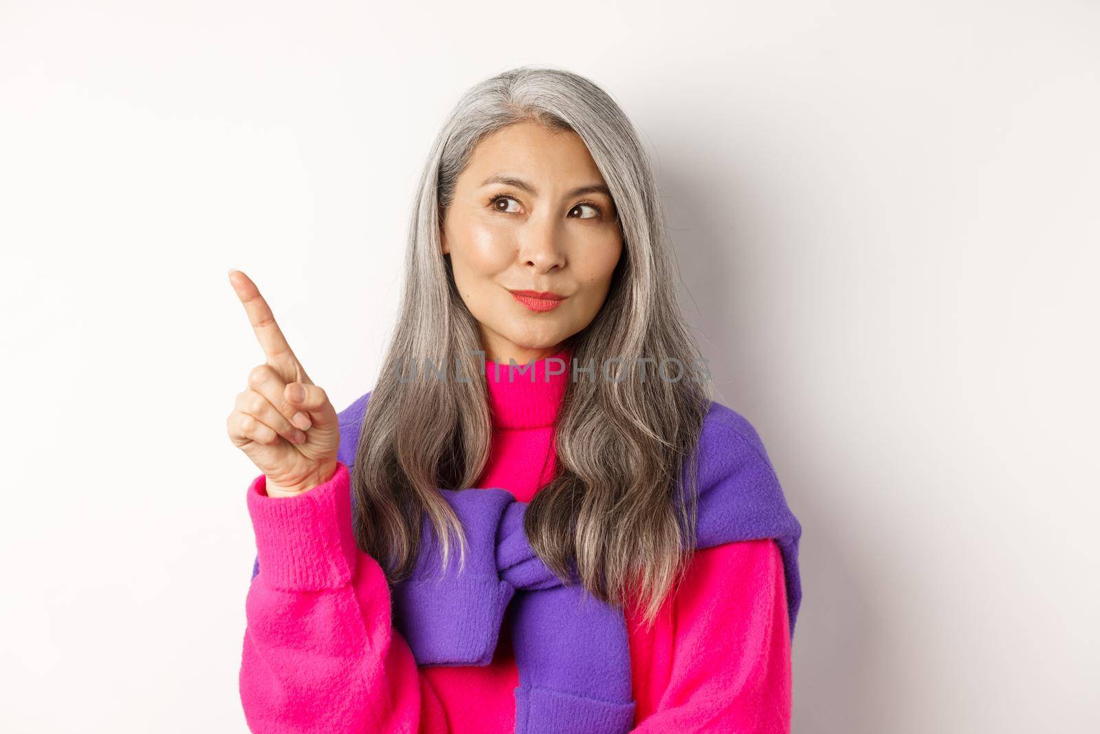 Close-up of stylish korean senior woman with grey hair looking and pointing upper left corner, showing promotion offer, standing over white background.