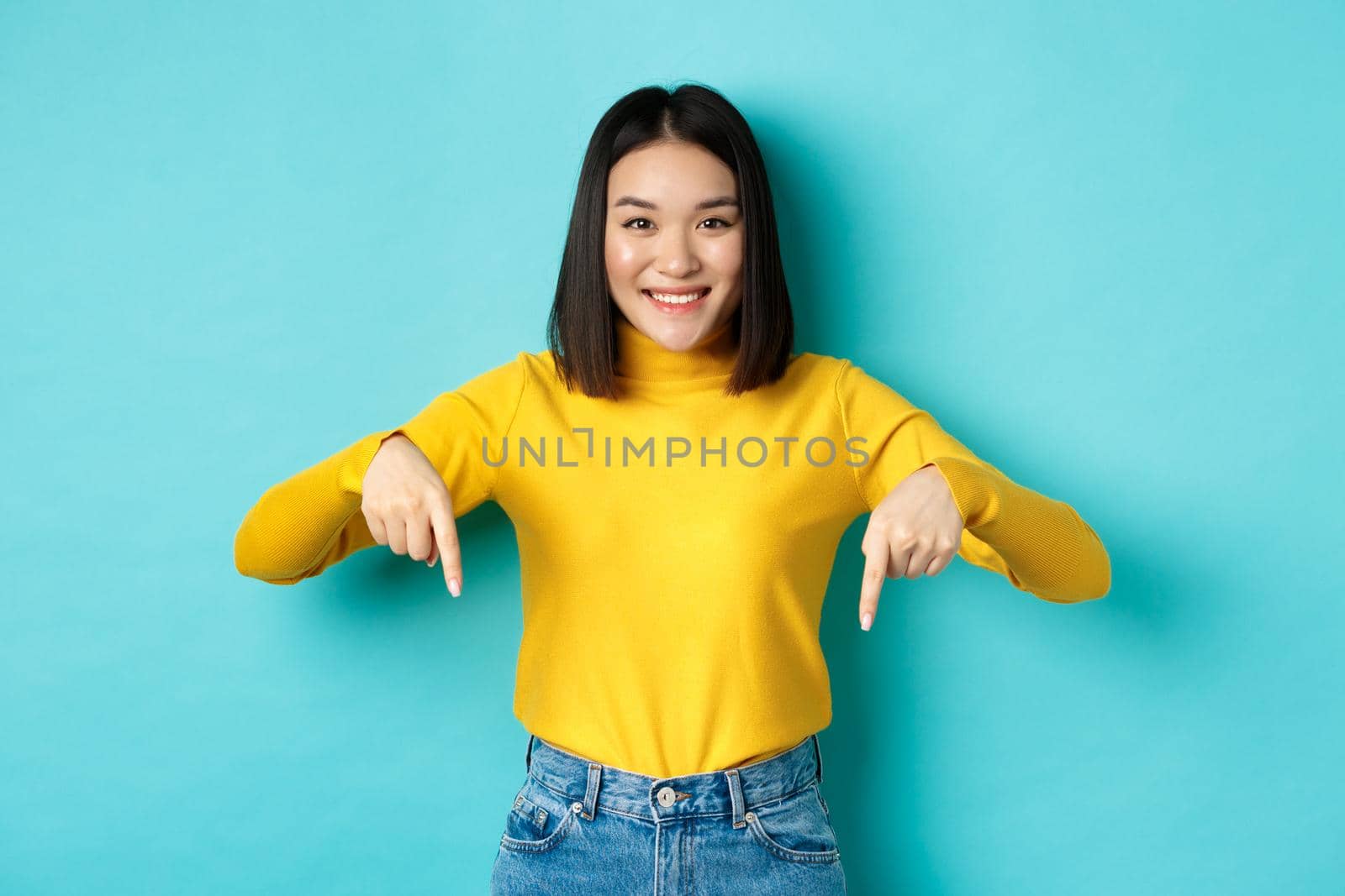 Shopping concept. Beautiful korean girl with happy smile, pointing fingers down at banner, standing against blue background.