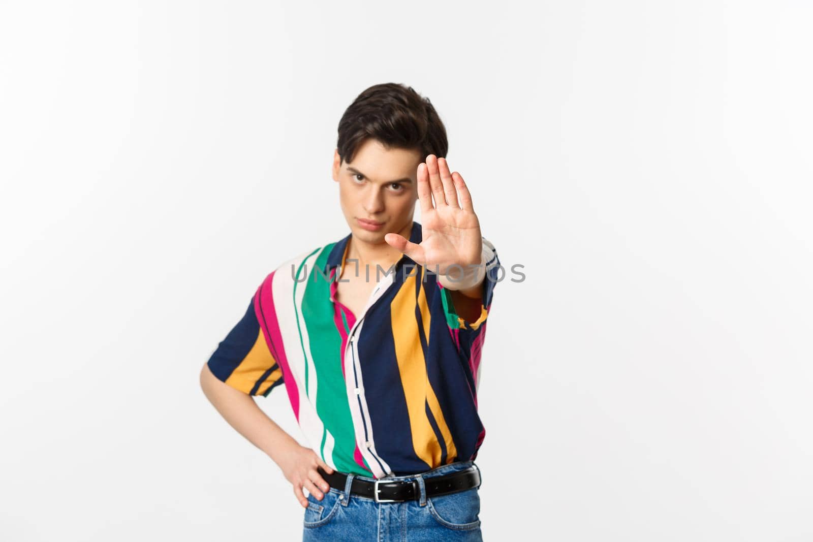 Serious young man extend hand, showing stop gesture, disapprove and prohibit action, standing over white background by Benzoix