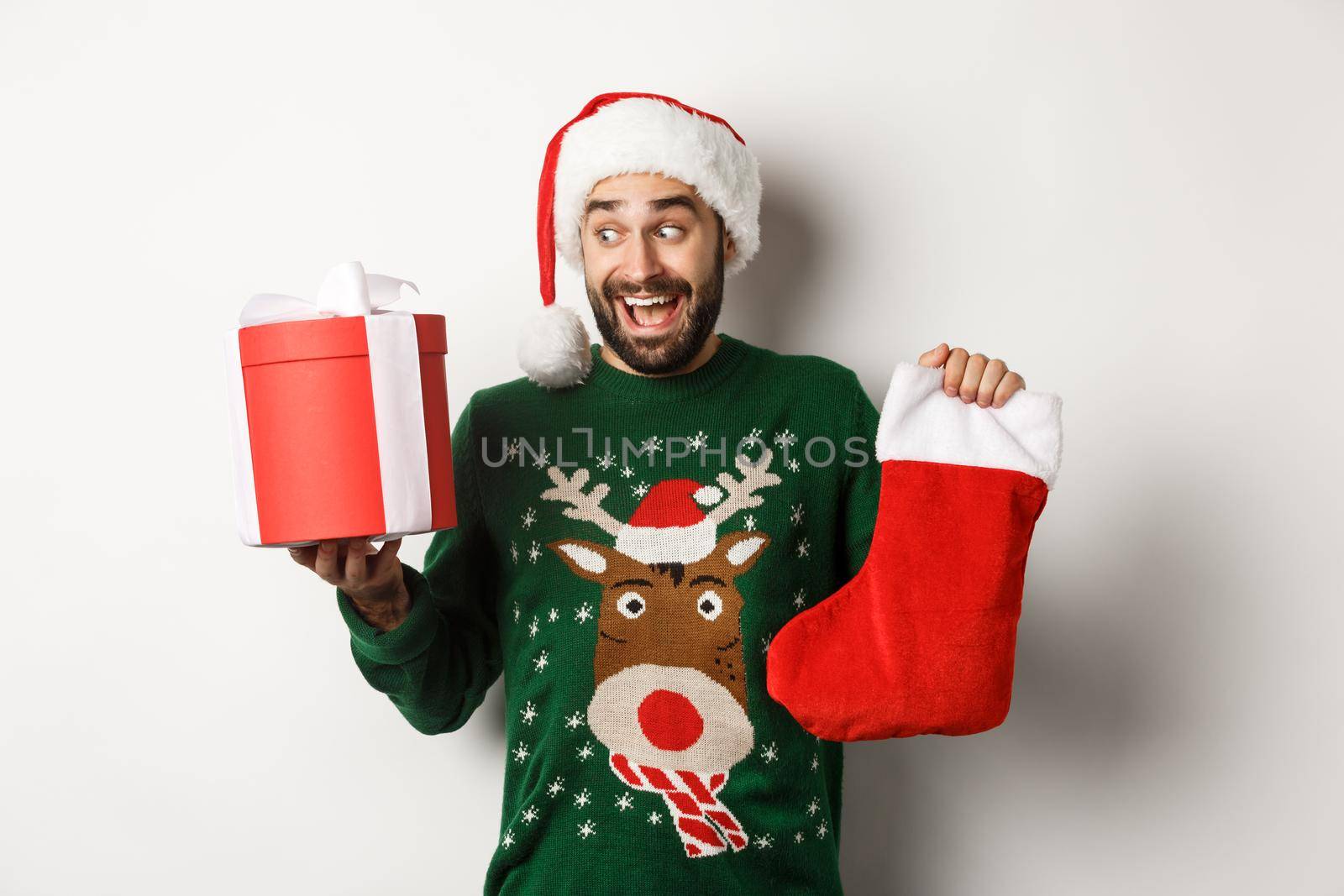 Xmas and winter holidays concept. Happy man got gifts on New Year eve, holding Christmas sock and present inside a box, standing over white background.