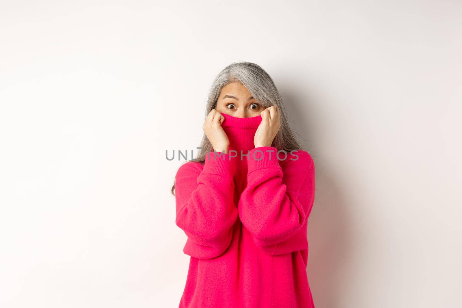 Portrait of funny asian grandmother hiding face in sweater collar, peeking at camera silly, standing over white background.