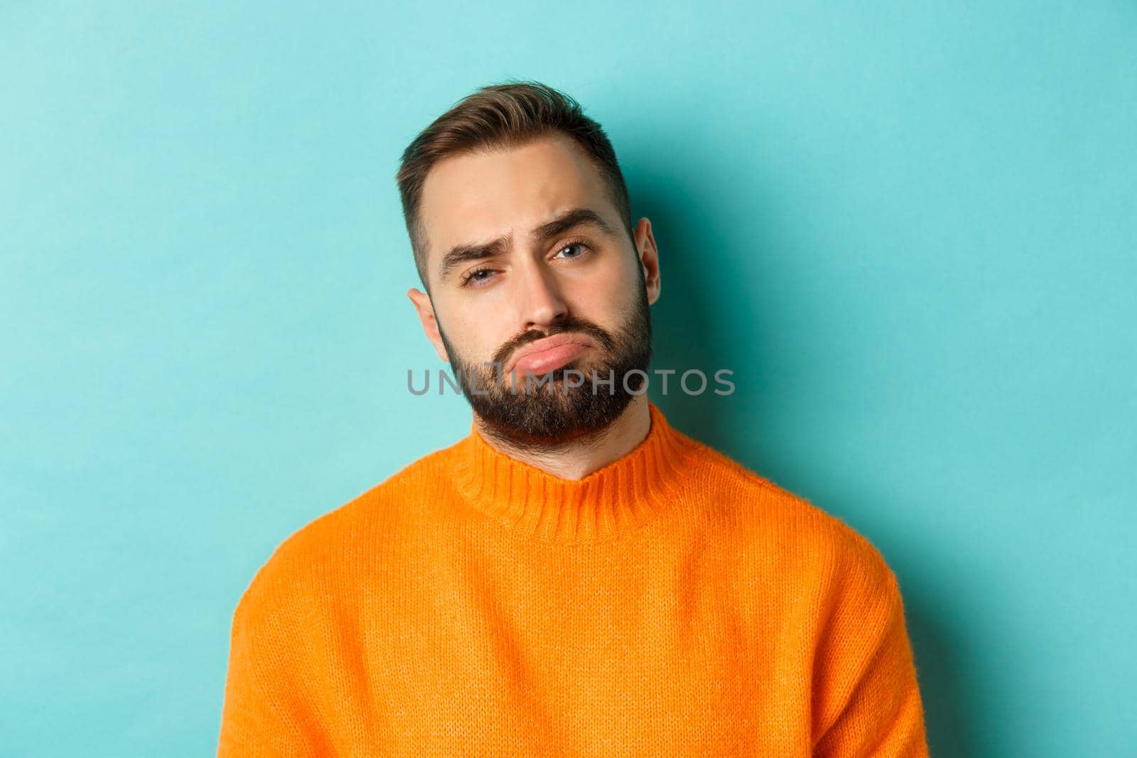 Close-up of upset young man sulking, looking disappointed and distressed, standing in orange sweater against turquoise background.
