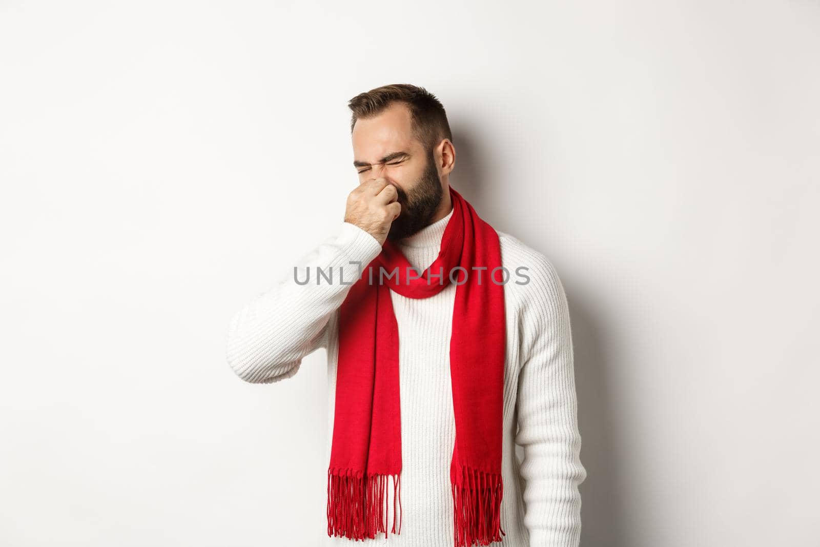 Displeased guy shut his nose from bad smell, grimacing disgusted, standing over white background in white sweater with red scarf.