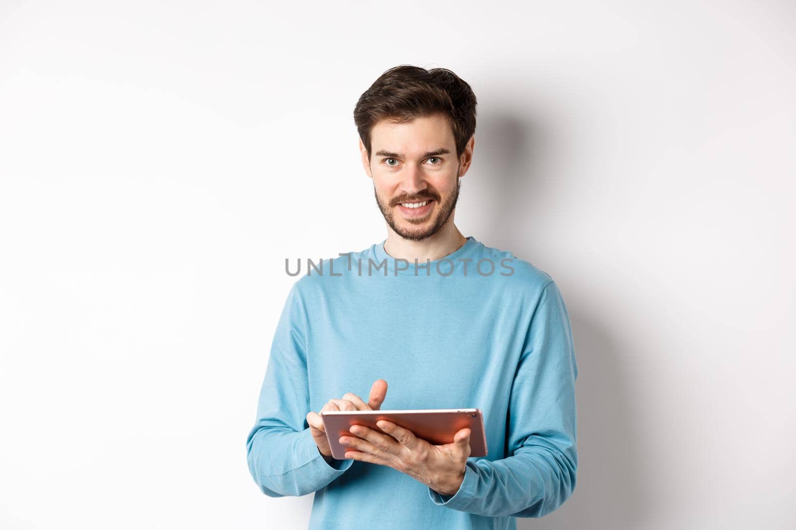 E-commerce. Smiling caucasian man using digital tablet and looking at camera, standing on white background.