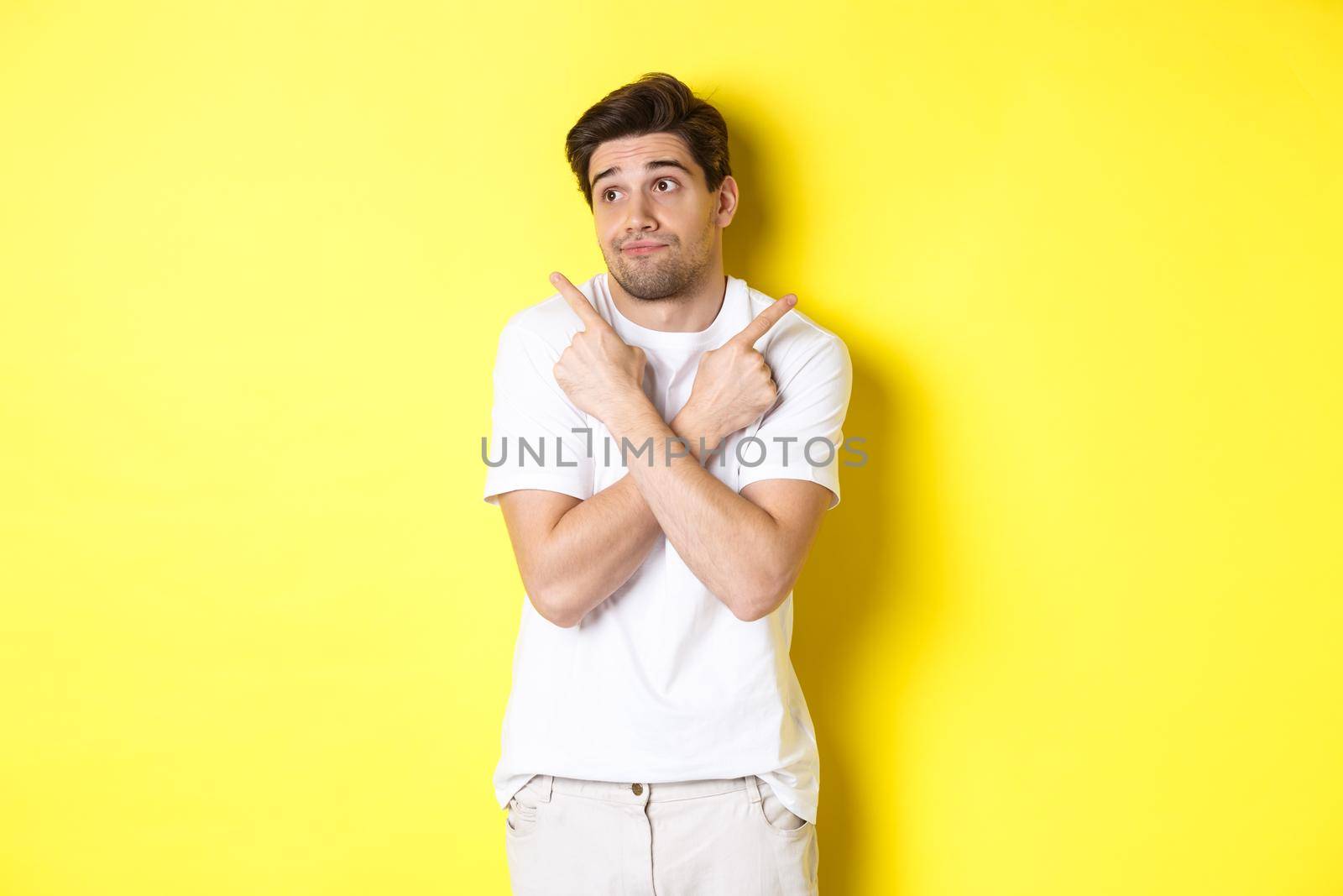 Indecisive man shrugging, pointing fingers sideways, troubled to choose, standing over yellow background.