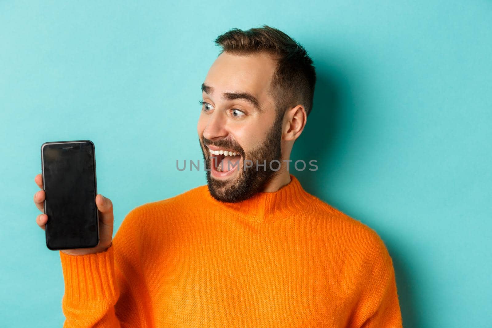 Close-up of young bearded man showing phone screen and looking amazed, wearing orange sweater, standing against studio background by Benzoix