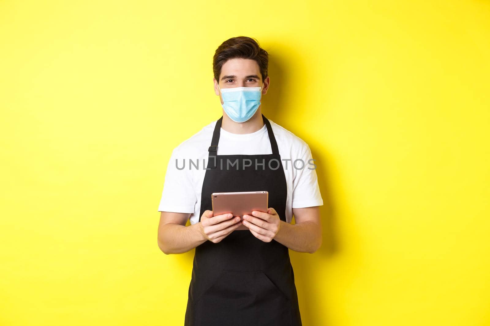 Concept of covid-19, small business and pandemic. Waiter in black apron and medical mask taking order, holding digital tablet, standing over yellow background.
