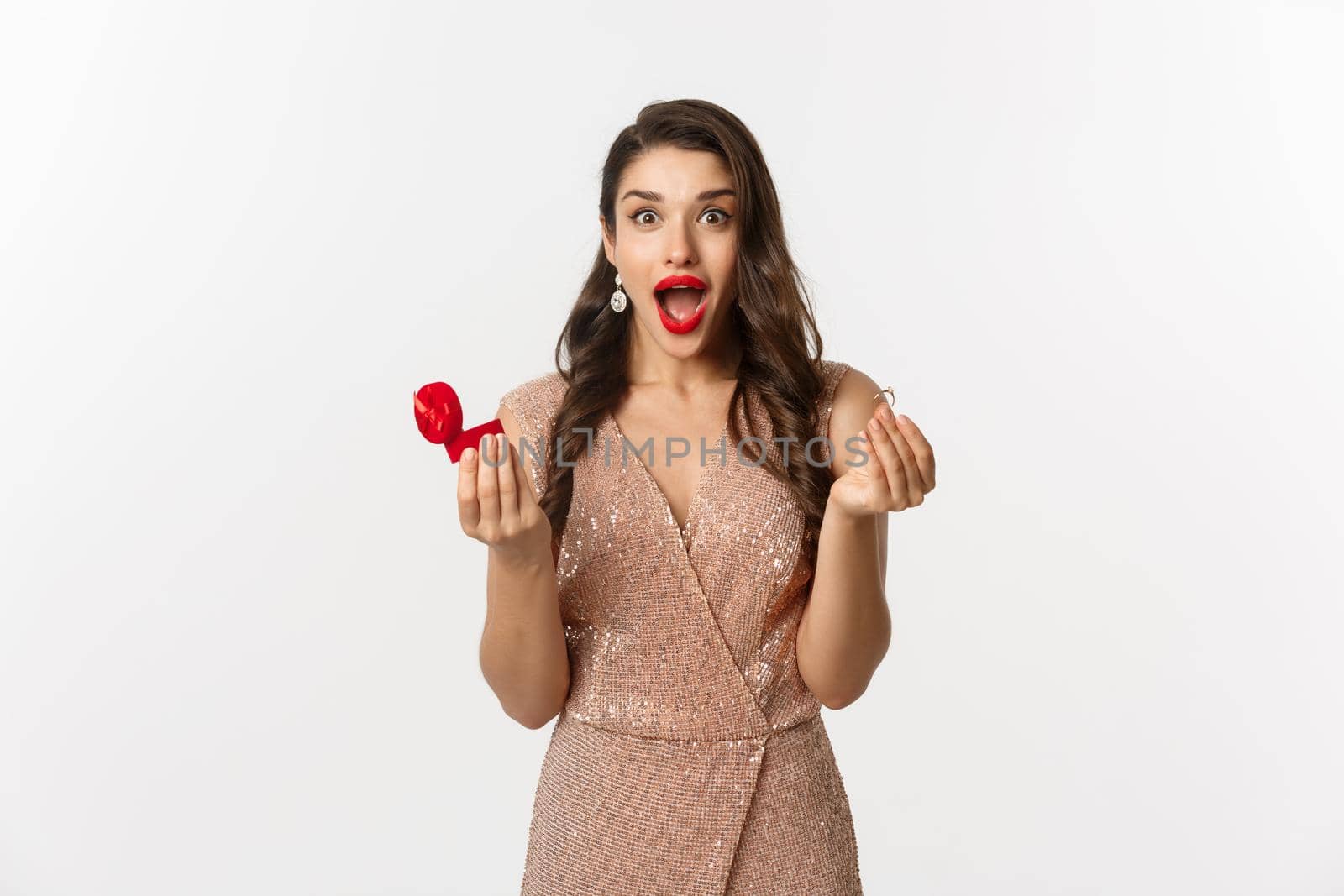 Excited woman holding engagement ring and red box, looking surprised and amazed, receive marriage proposal on date, wearing elegant dress, standing over white background by Benzoix