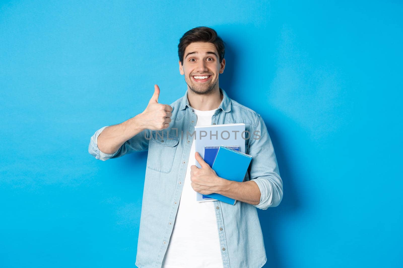 Happy young male model holding notebooks and showing thumb-up pleased, smiling and recommending courses, standing over blue background.