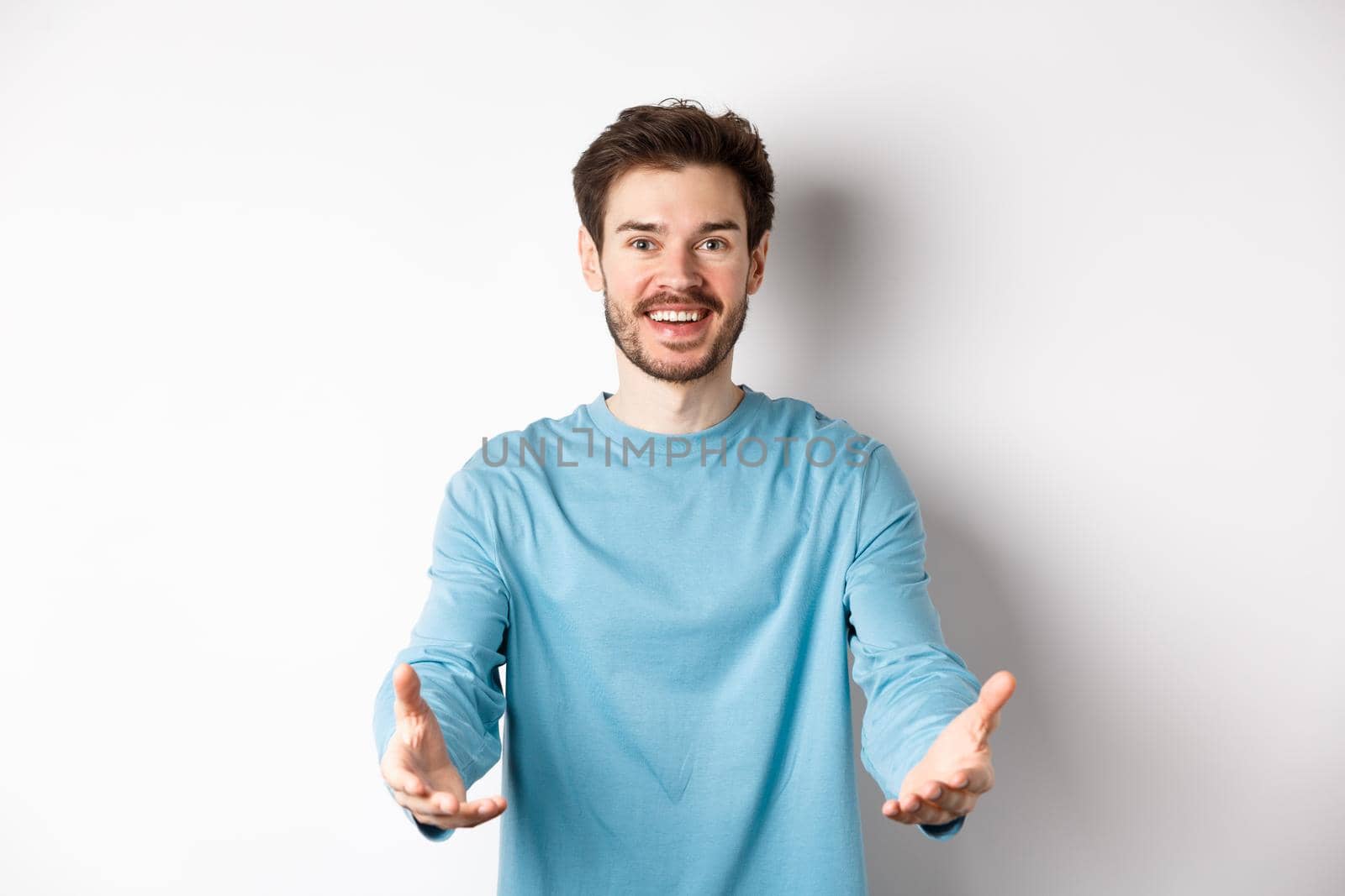 Attractive caucasian guy in casual clothes stretch out hands to hold something, receiving and smiling, standing over white background.