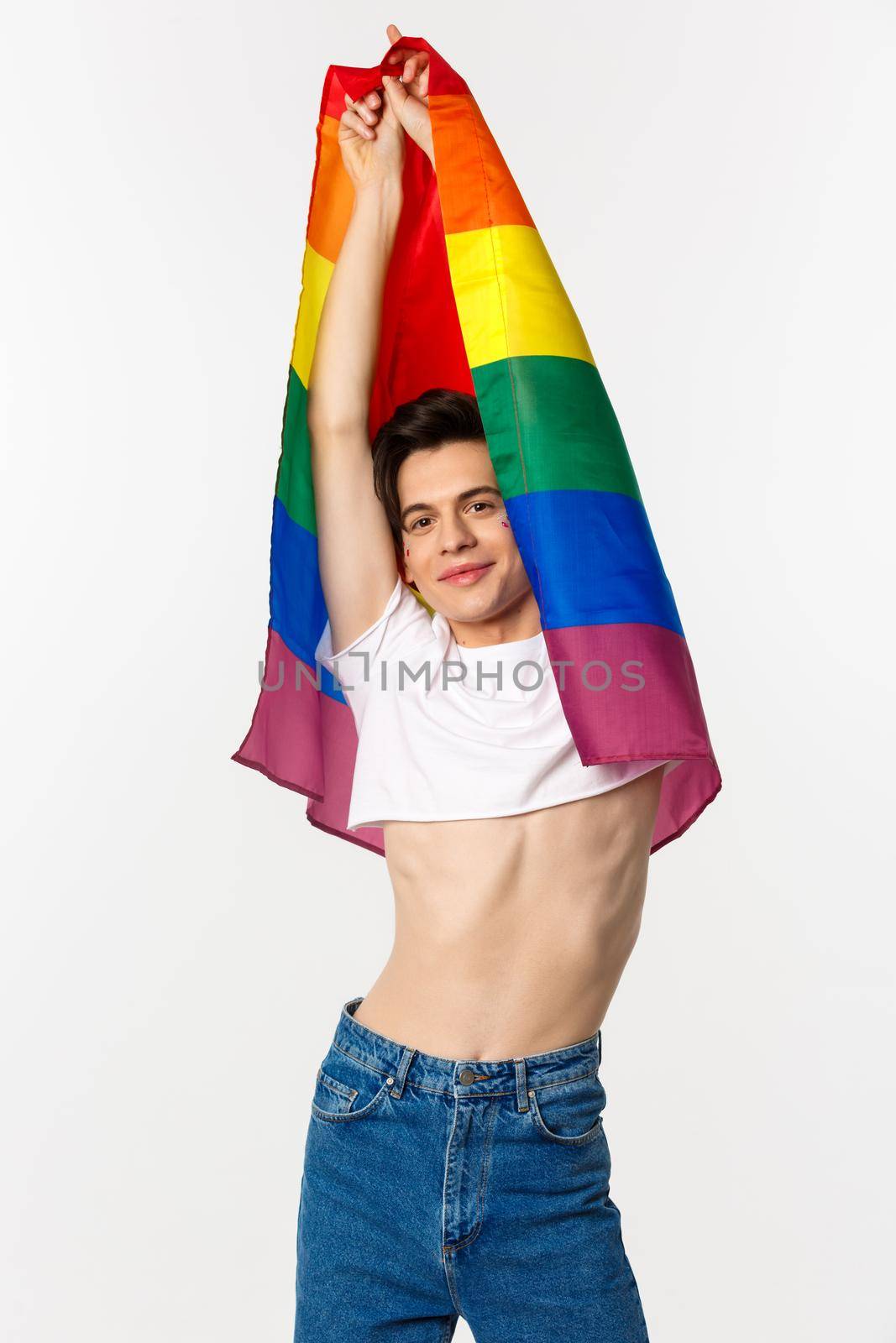 Vertical view of beautiful androgynous gay man raising rainbow flag and smiling happy, standing in crop top and jeans against white background by Benzoix