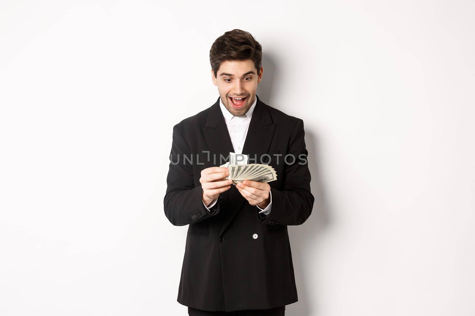 Image of excited handsome businessman, counting money and smiling amused, standing against white background in suit.