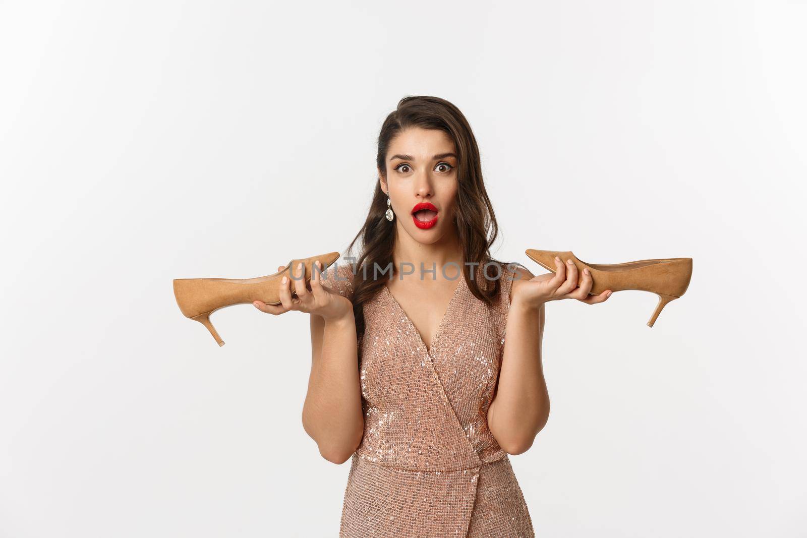 Party and celebration concept. Attractive woman in elegant dress holding pair of heels and looking surprised, dressing up for Christmas, white background.