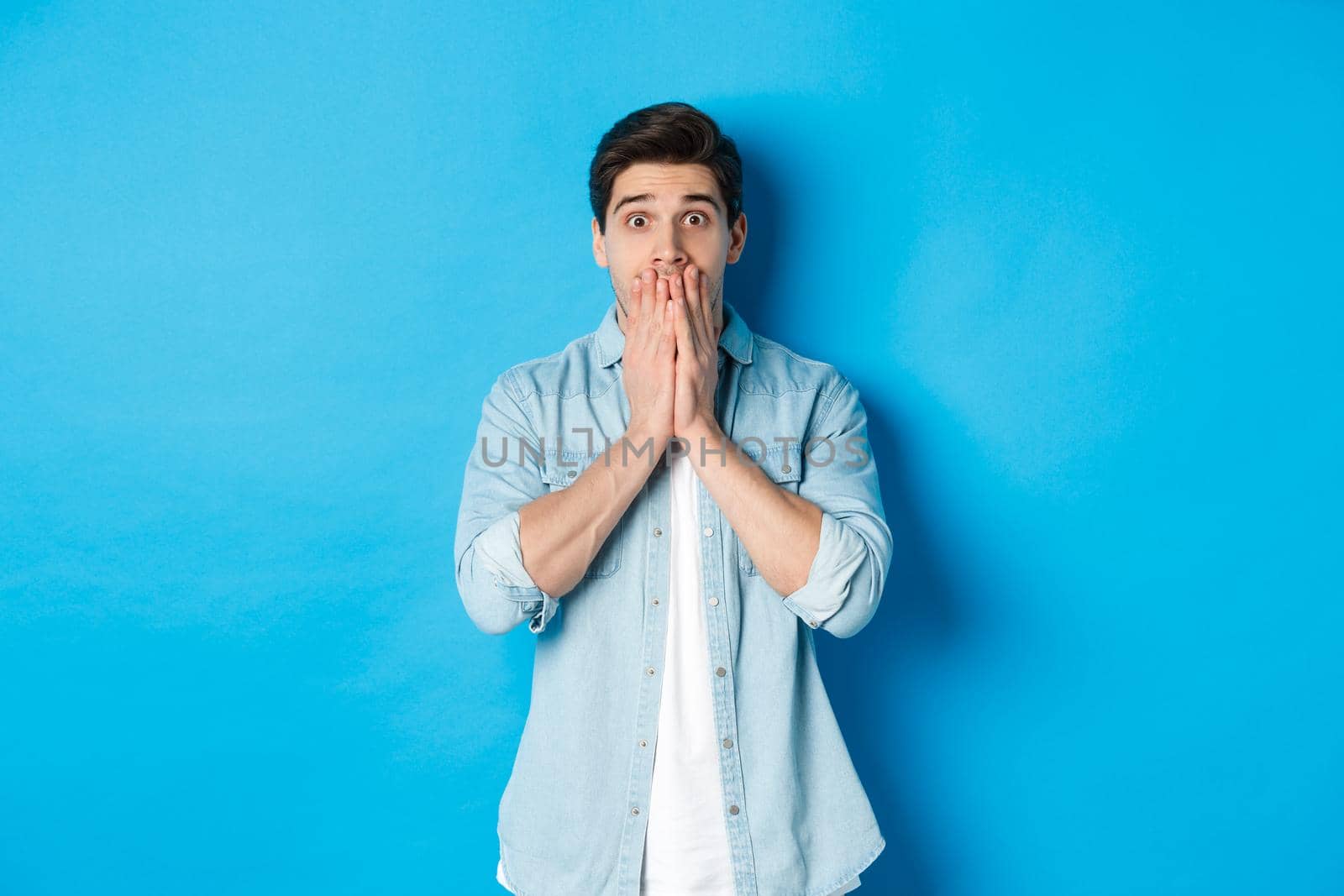 Portrait of shocked adult man looking at camera in awe, gasping and covering mouth with hands, standing over blue background by Benzoix