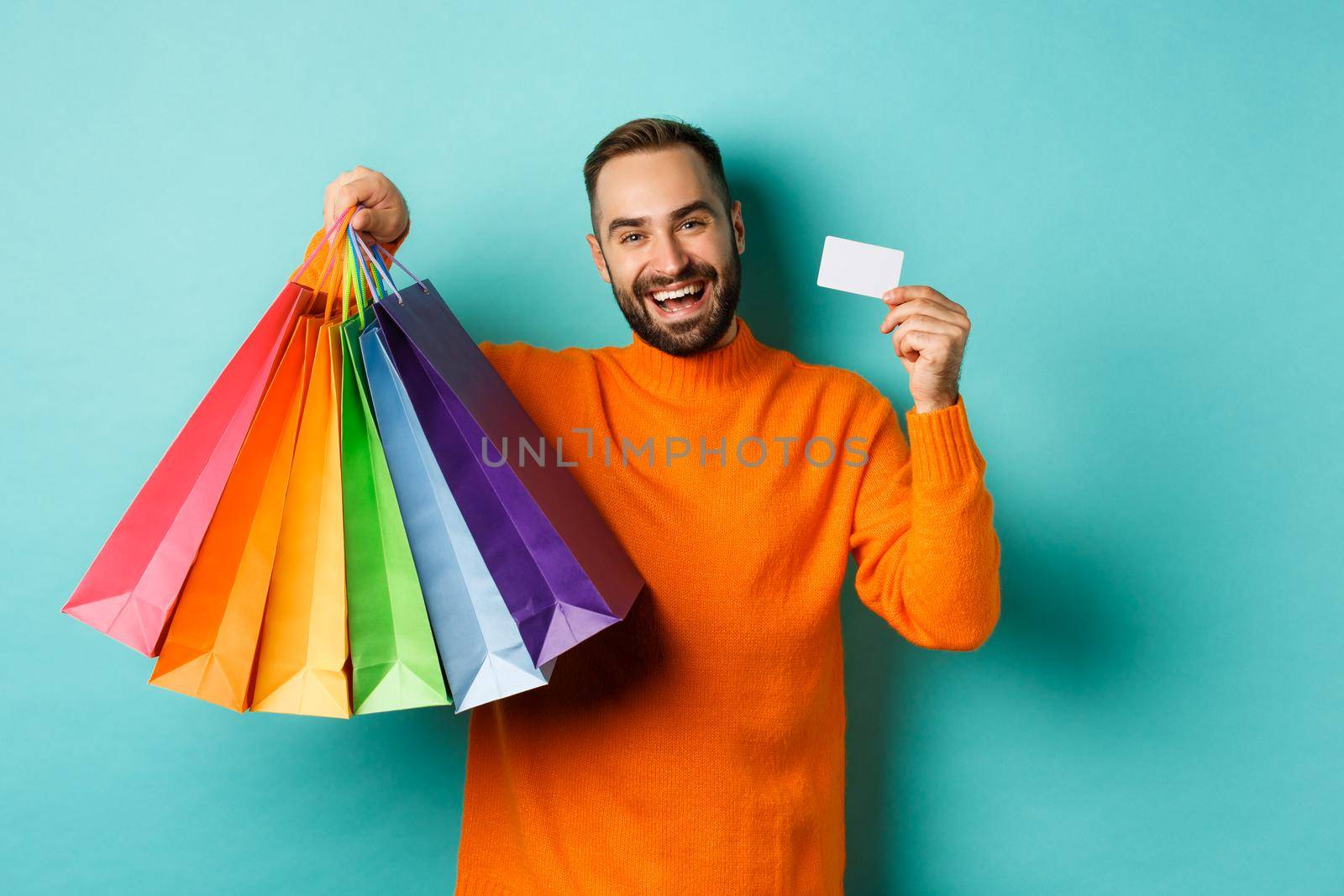 Happy aduly man showing credit card and shopping bags, standing against turquoise background. Copy space