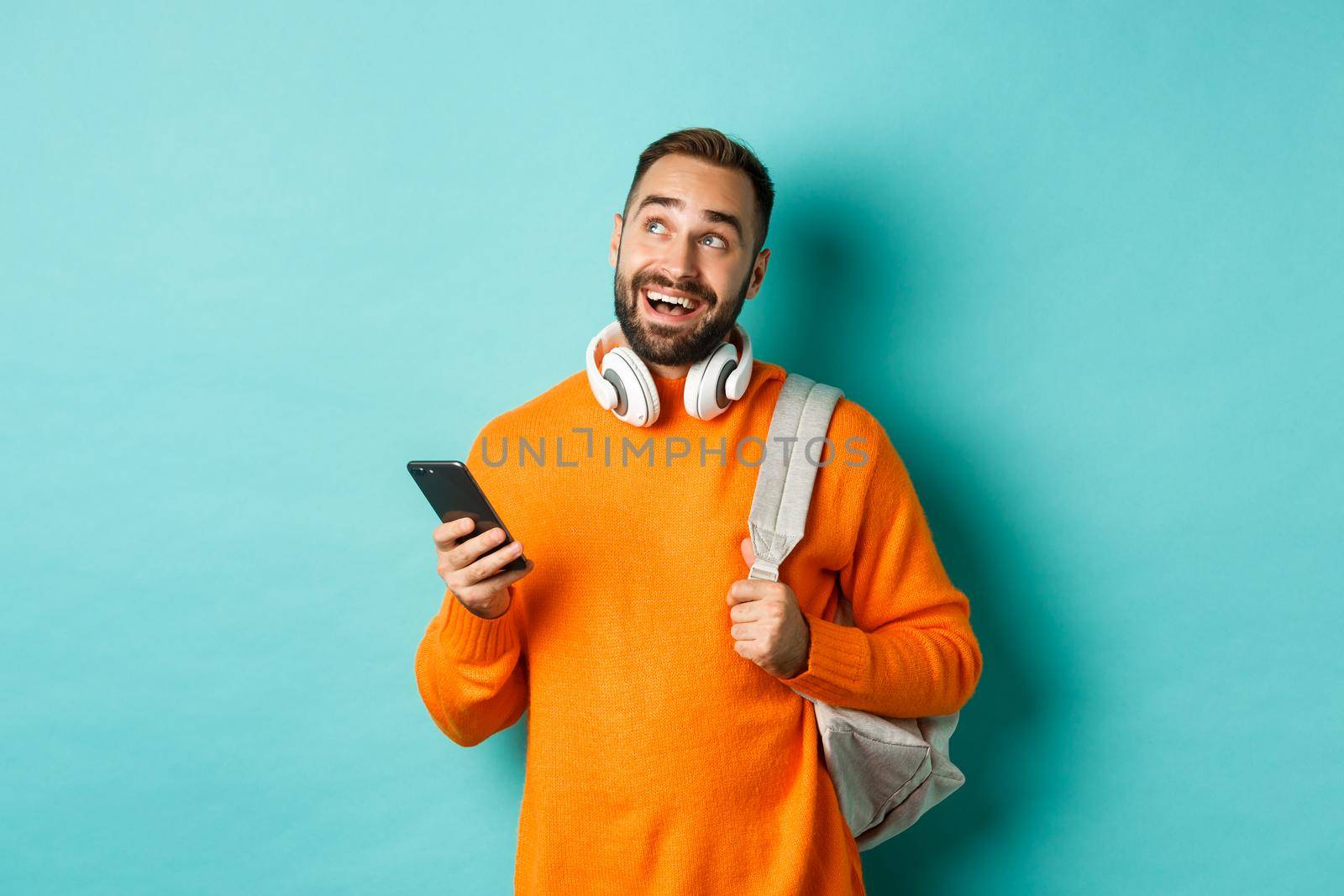 Image of handsome young man with backpack and headphones, imaging or thinking while using smartphone, standing over light blue background.