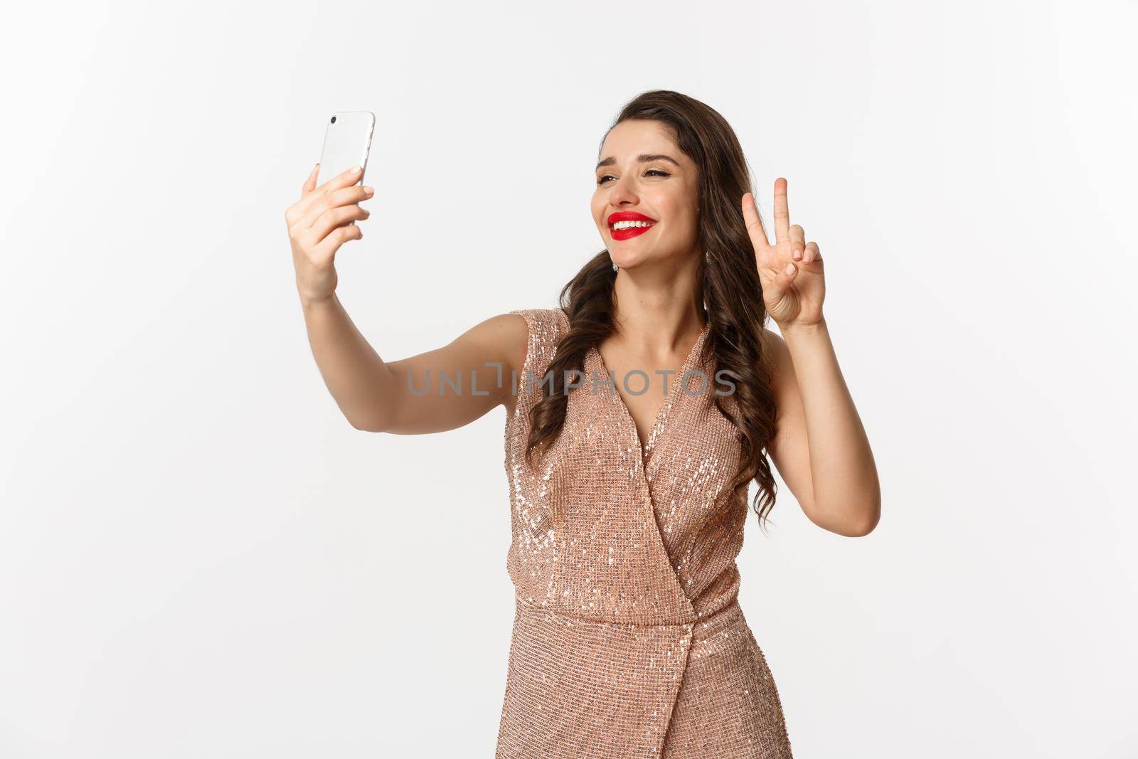 Attractive woman in luxury dress showing peace sign at phone camera, taking selfie on smartphone, posing in christmas party outfit, standing over white background by Benzoix
