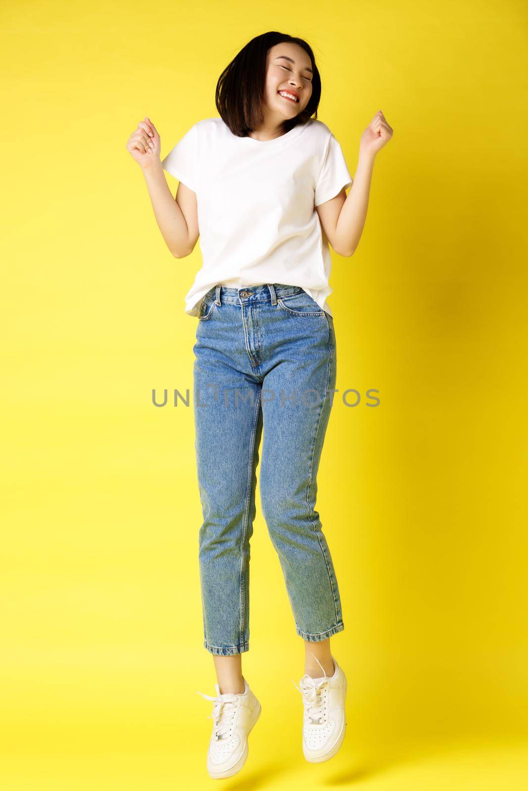 Full size shot of carefree asian woman jumping and dancing, having fun, posing in jeans and white t-shirt over yellow background by Benzoix
